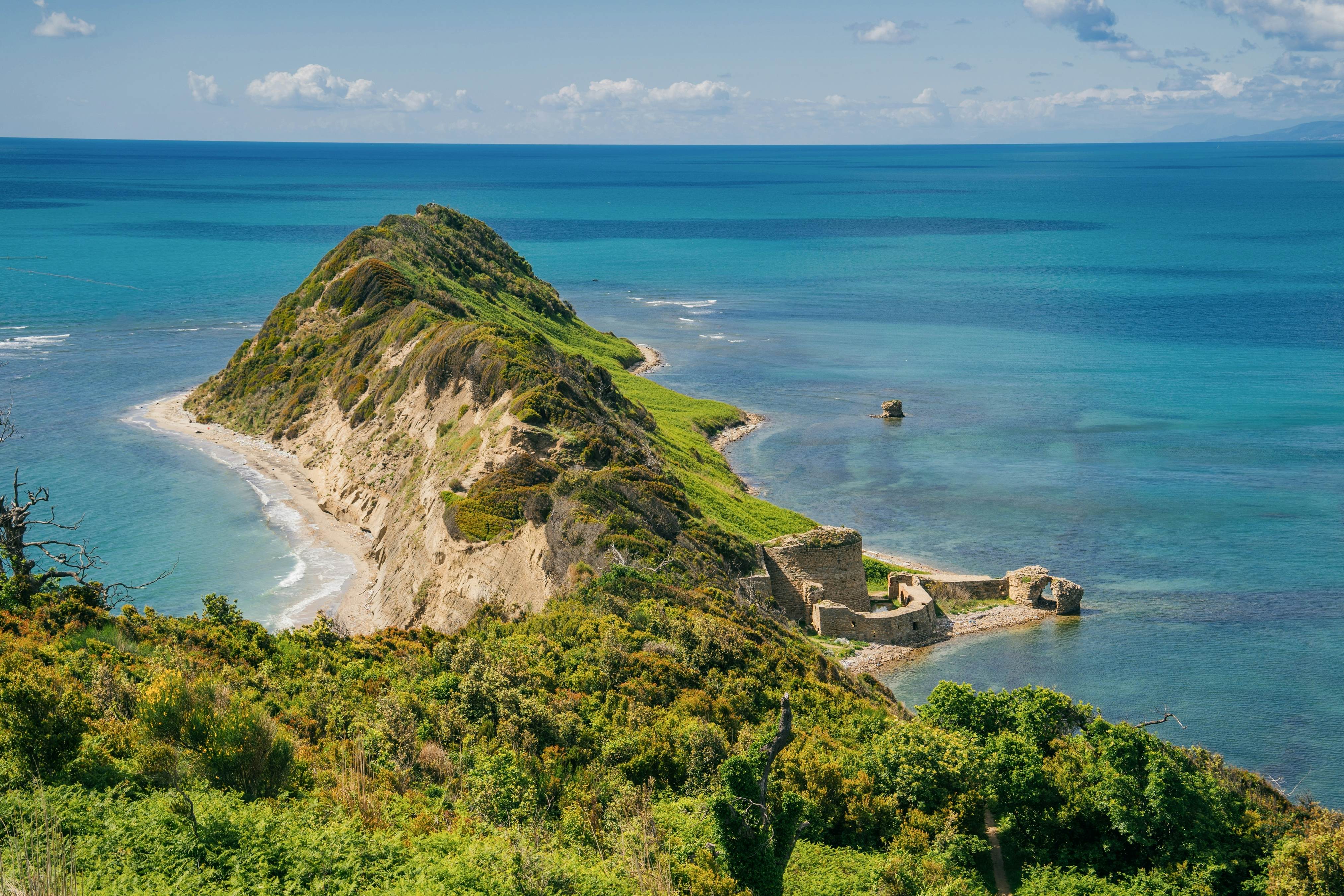 A view from above of a green cape jutting into the sea. Beaches and a ruined fortress sit along the shore of the hilly headland.