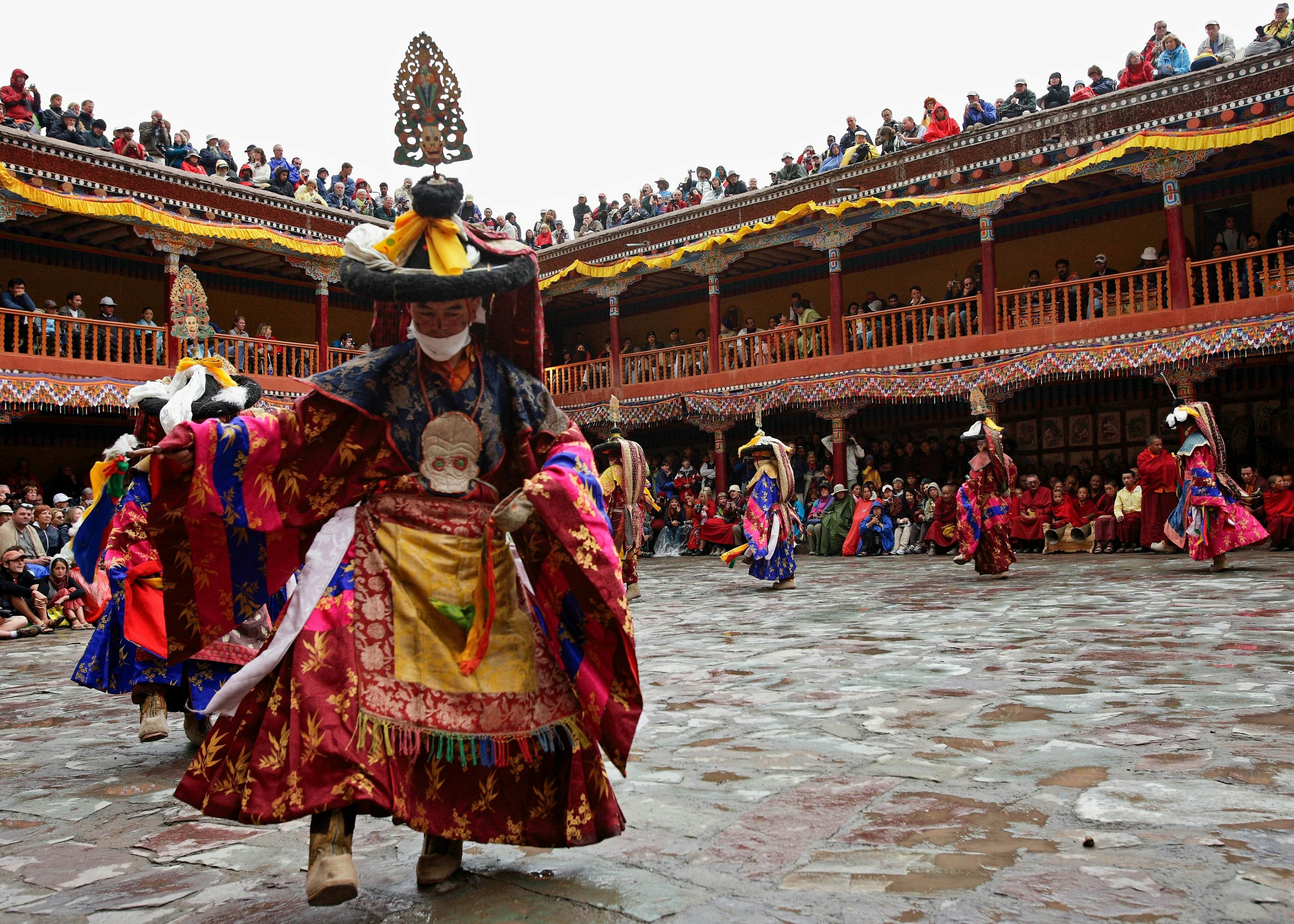 Monks in colorful costumes and elaborate headdresses perform a ritual dance in the courtyard of a monastery as people watch from the balcony and roof.