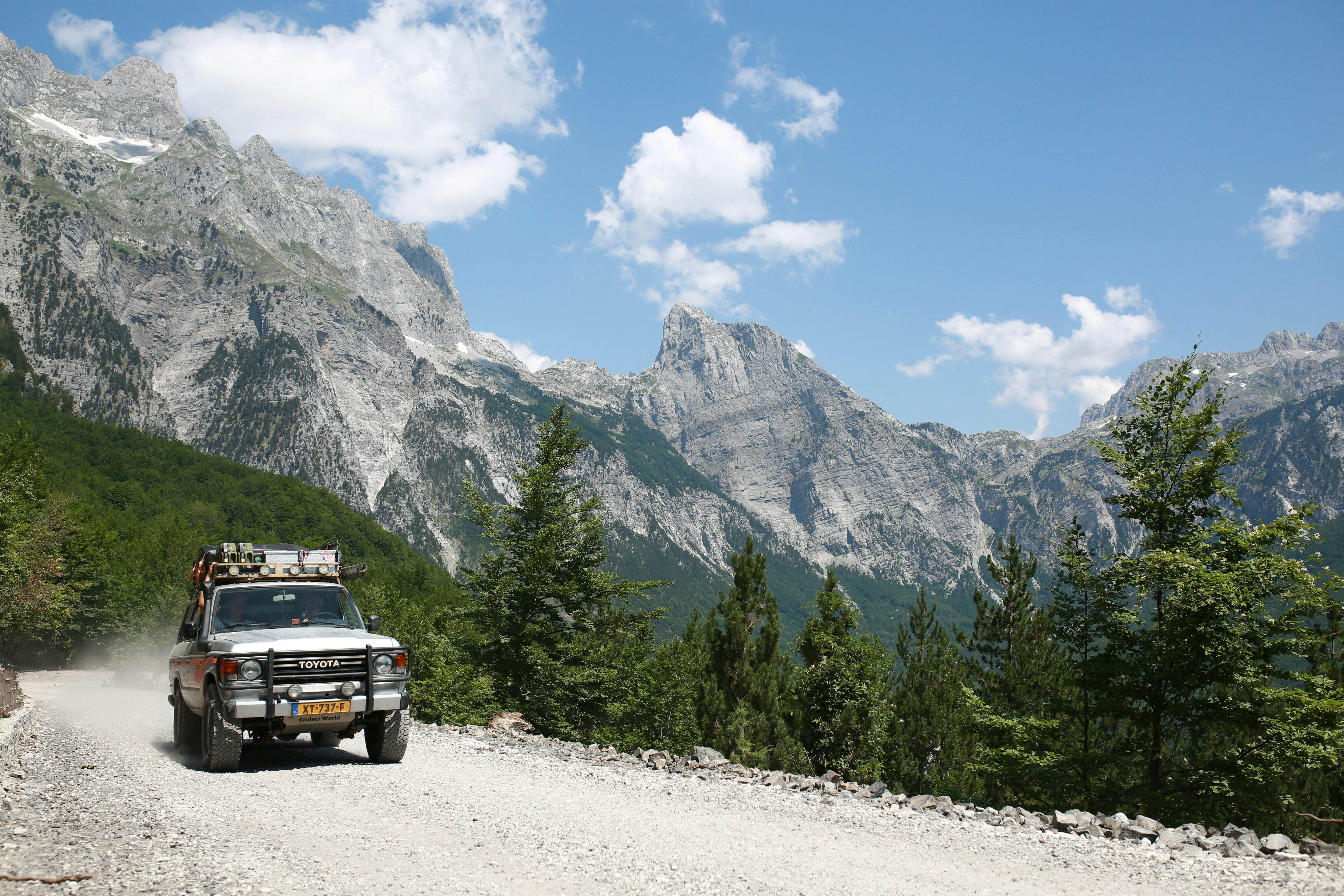 An SUV with large wheels and crates strapped to its roof drives on a dirt road through the mountains. Rocky peaks and ridges are visible in the distance.