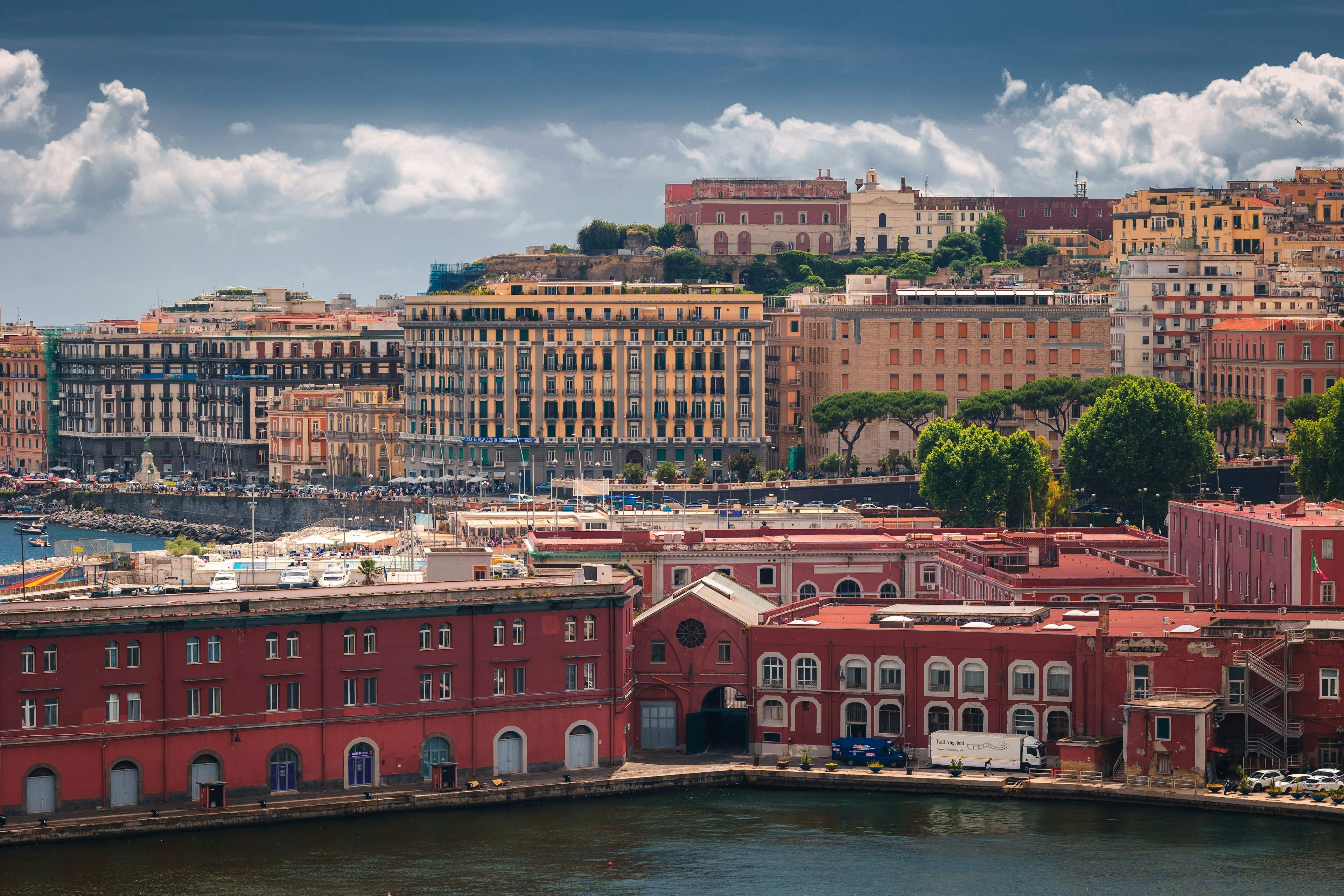 View of Naples' harbor, illuminated by soft, golden light. A variety of ships and boats are docked along the waterfront.