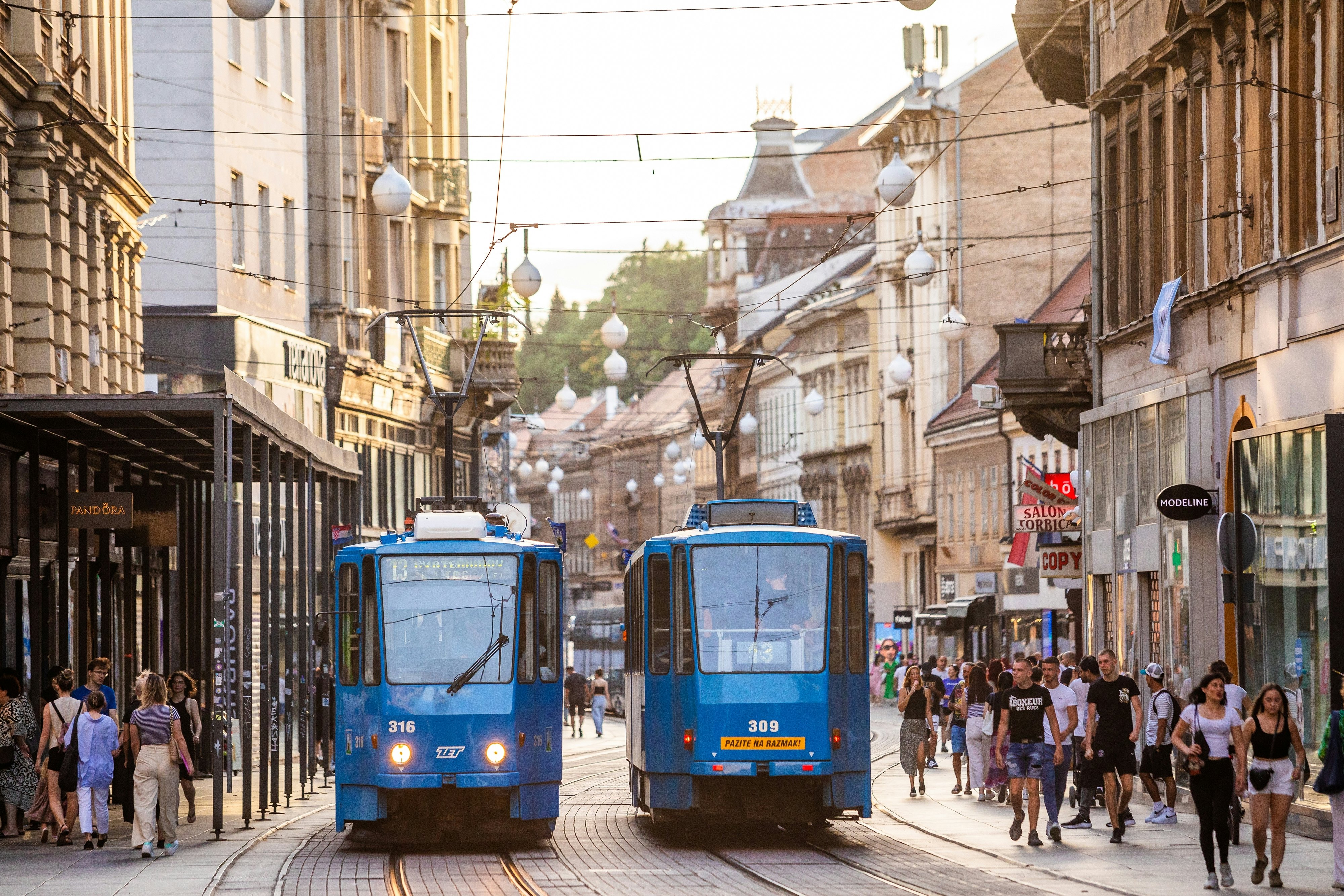 Two blue trams pass one another on a crowded street in a city.