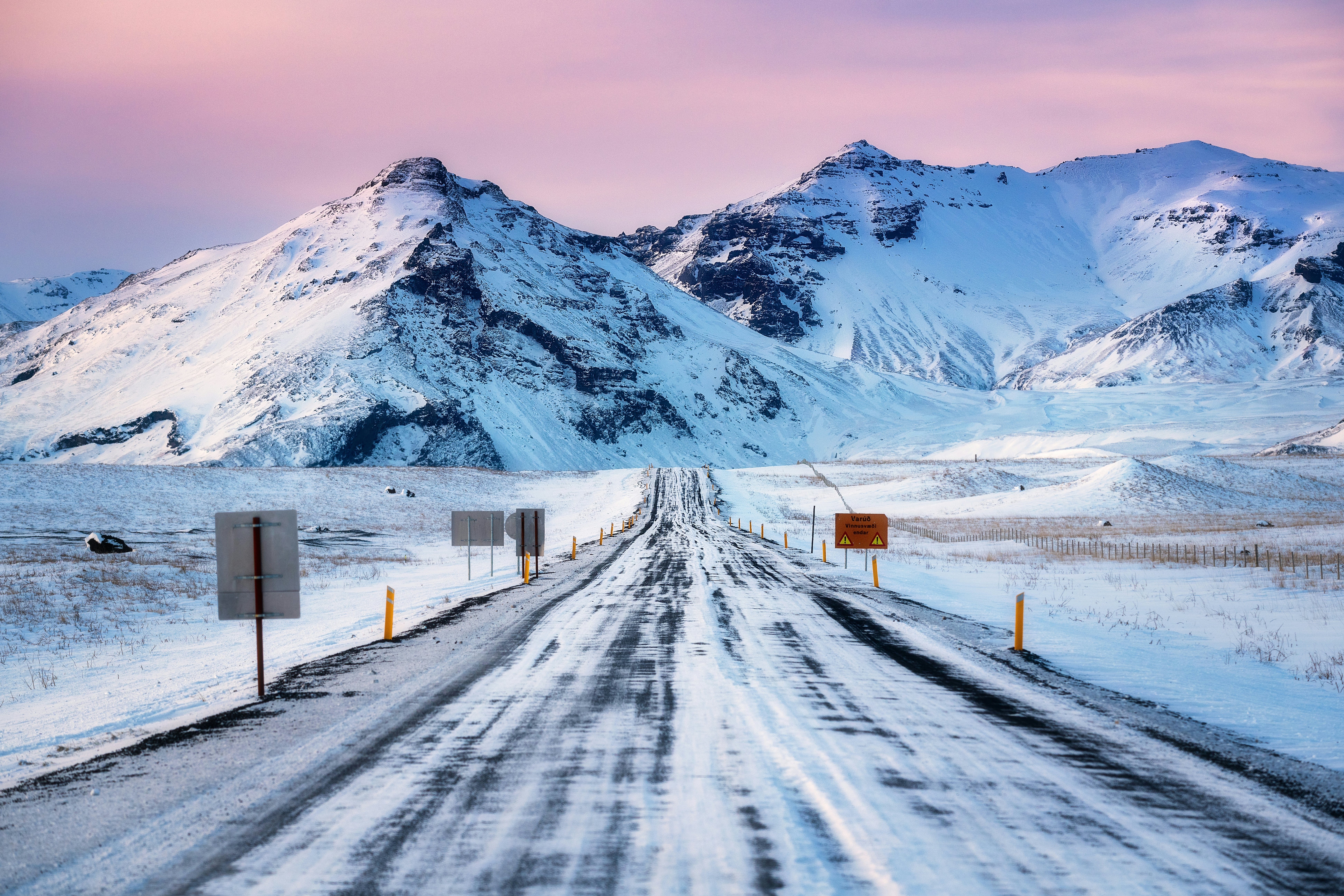 Scenic view from Ring Road Route 1 in Iceland during winter at sunset, with pink clouds, road signs, poles on the side of road, mountains in the background, driving through the beautiful Iceland