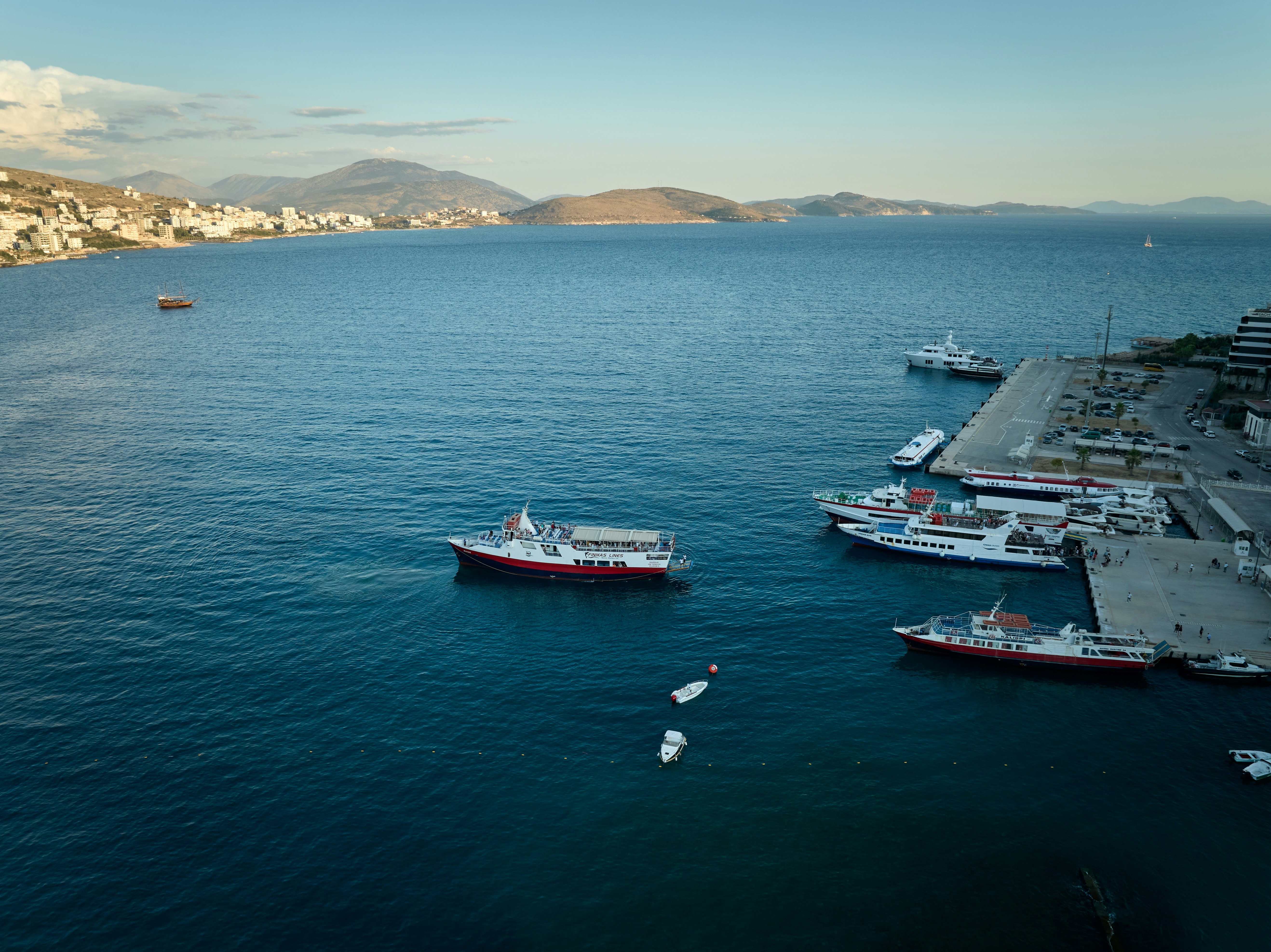 An aerial view of a ferryboat pulling out from a port. Hills and white-washed buildings are visible on the distant shore.