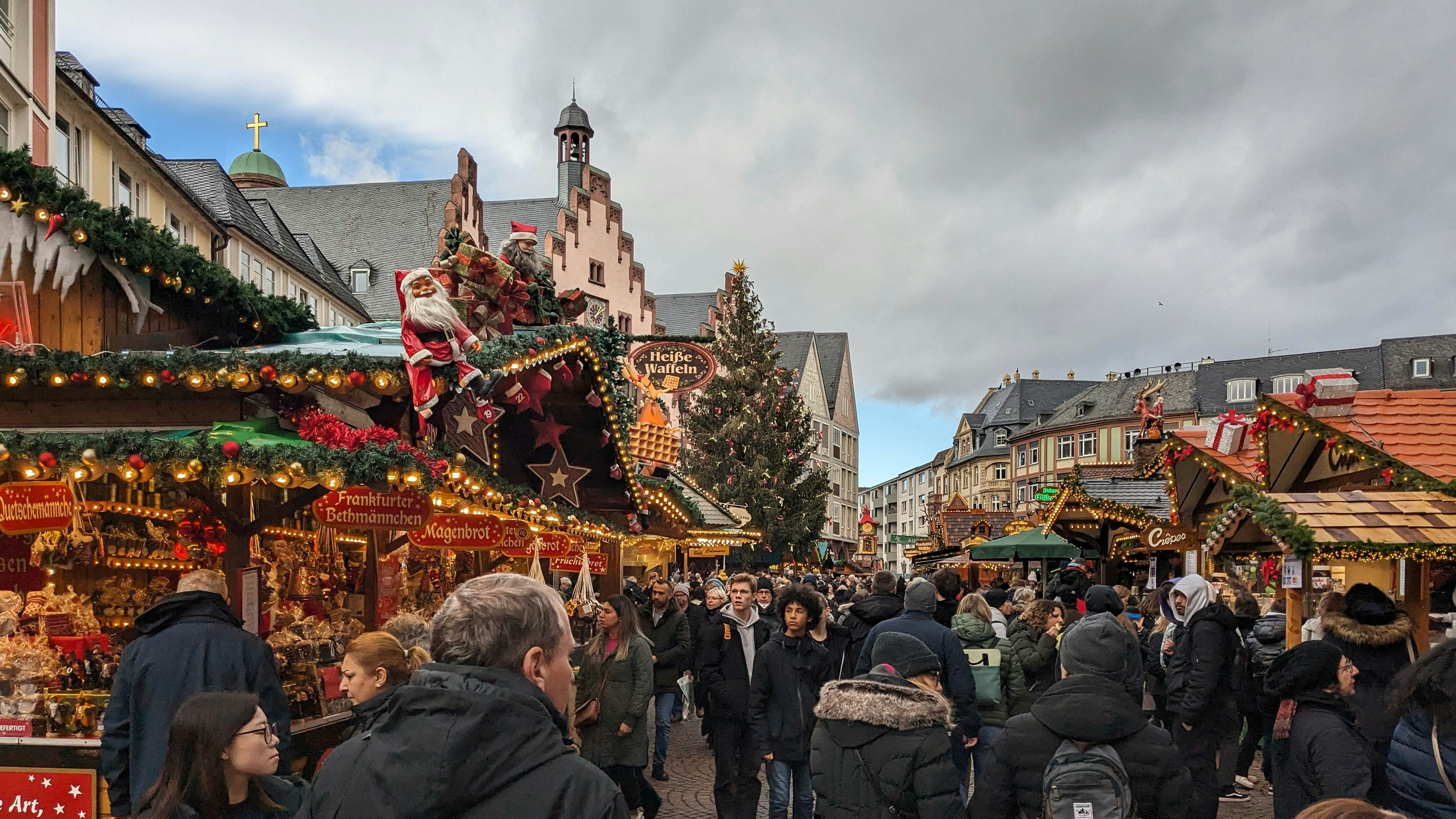 Frankfurt, Hesse, Germany - December 20, 2023: Traditional Christmas market with illuminated Bethmannchen stand, Santa Claus figure, and crowd beneath historic buildings.