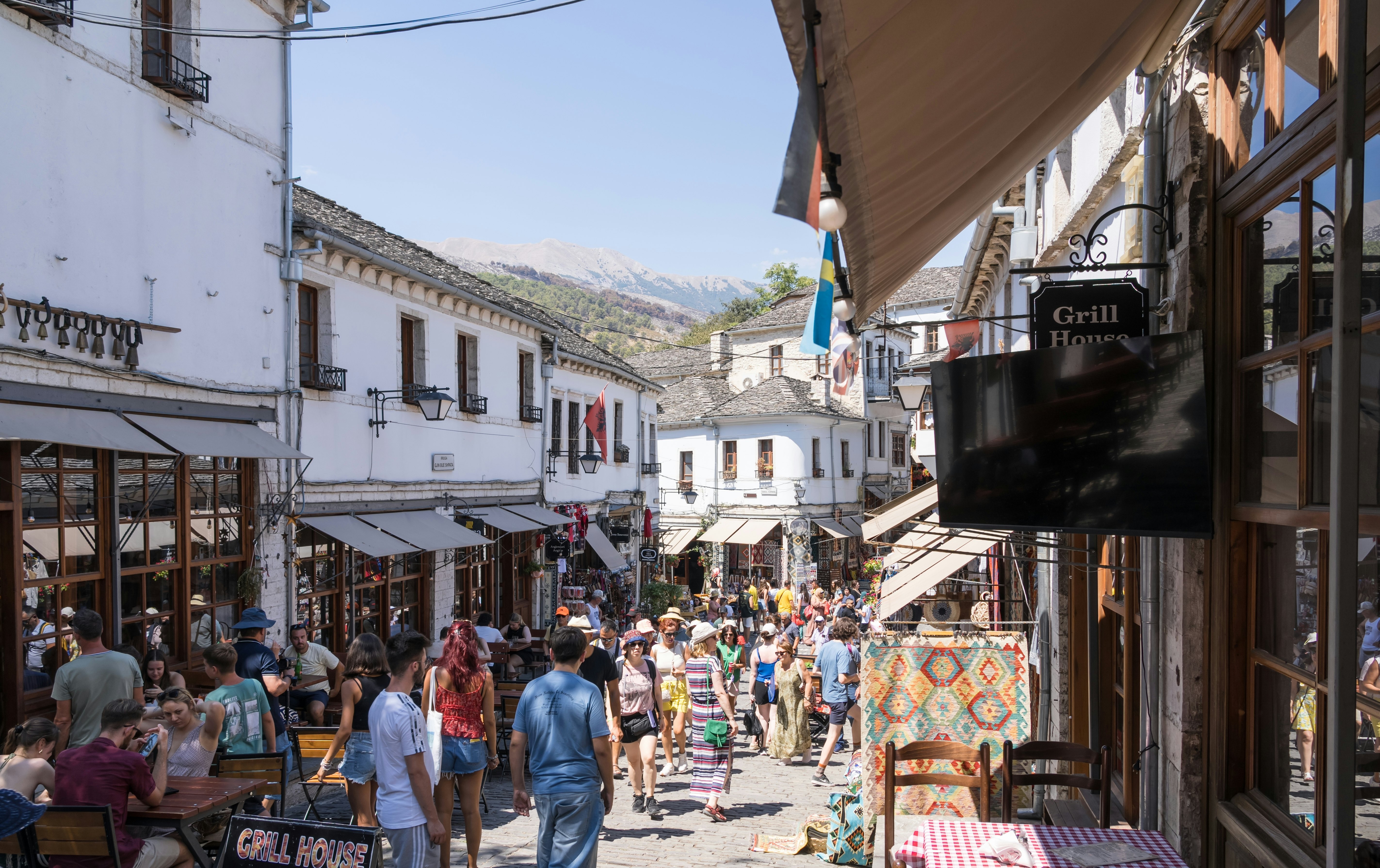 Crowds of people navigate an alley filled with shops and vendors in a historic mountain town filled with white-painted buildings.