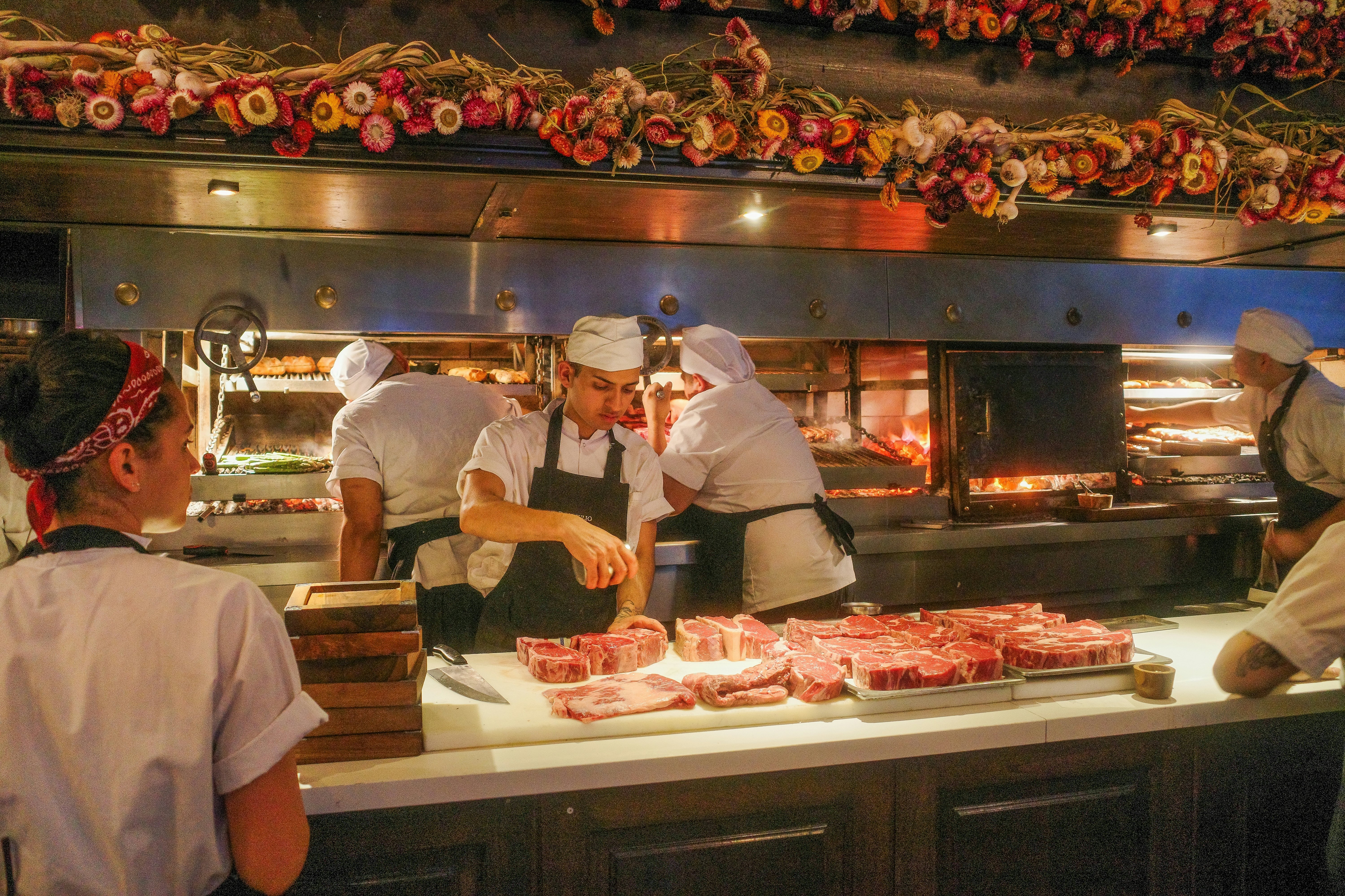 A chef seasons raw cuts of red meat on a counter at a restaurant. Other chefs and workers man the kitchen around him.