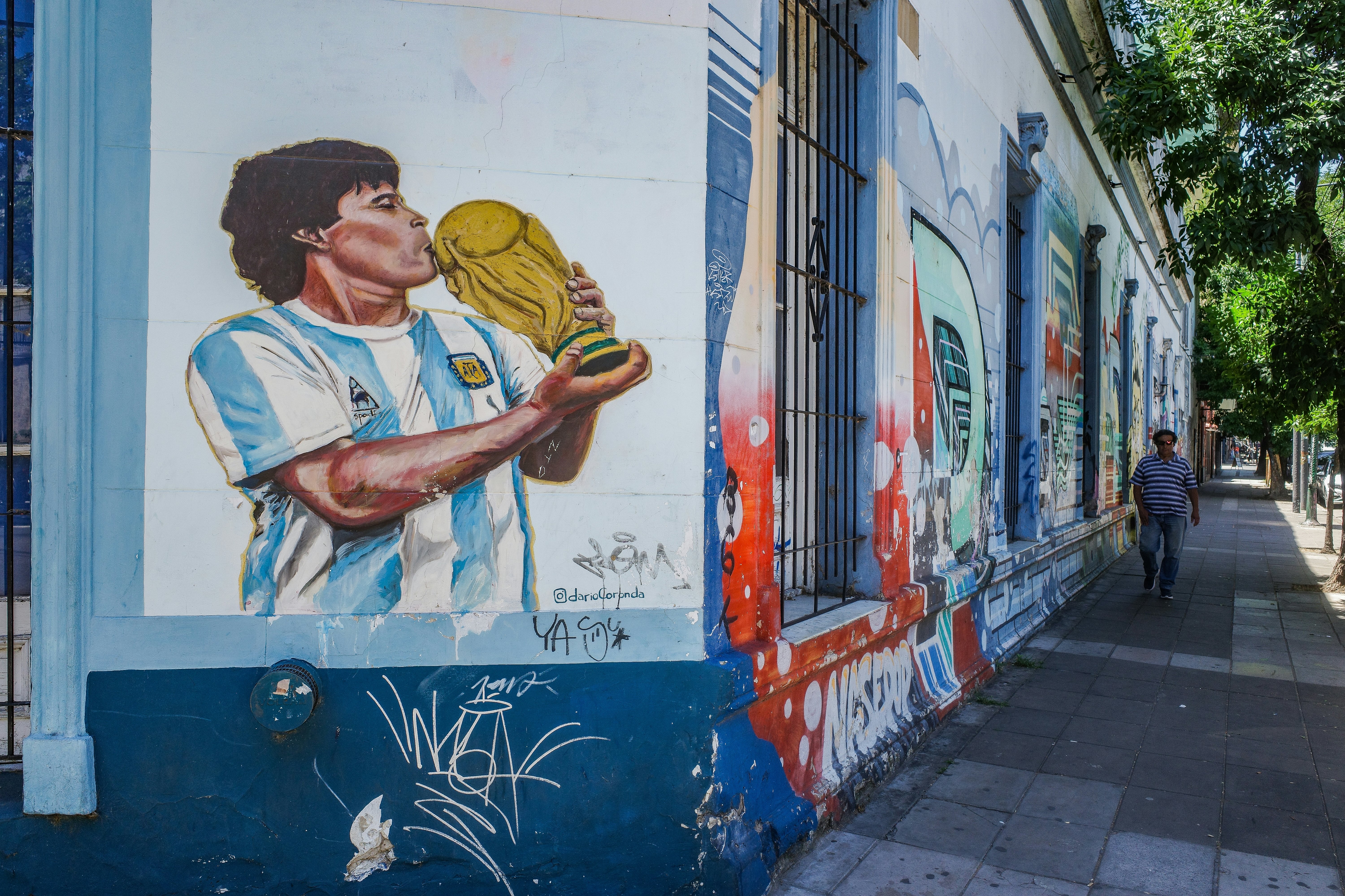 A mural of soccer player Diego Maradona kissing the World Cup trophy is painted on the corner of a building, with other murals visible around the corner.