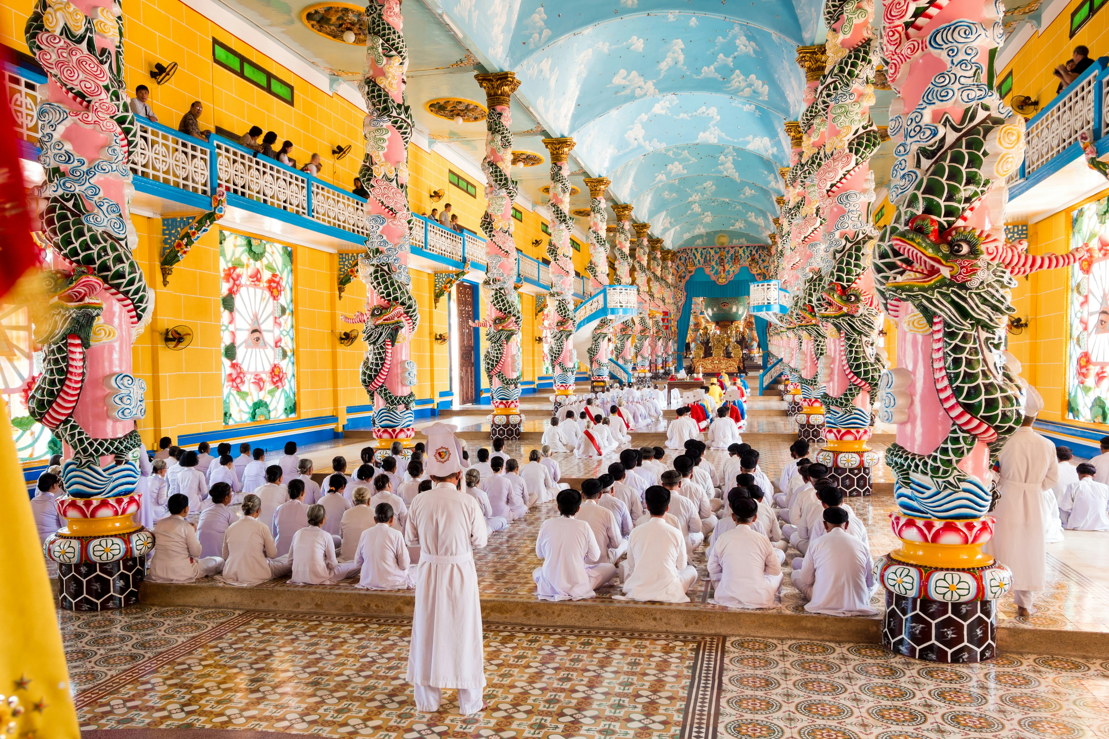 Participants in white robes sit in prayer in a sanctuary during a religious ritual. The sanctuary is painted yellow, with carved, painted dragons and other colorful details.