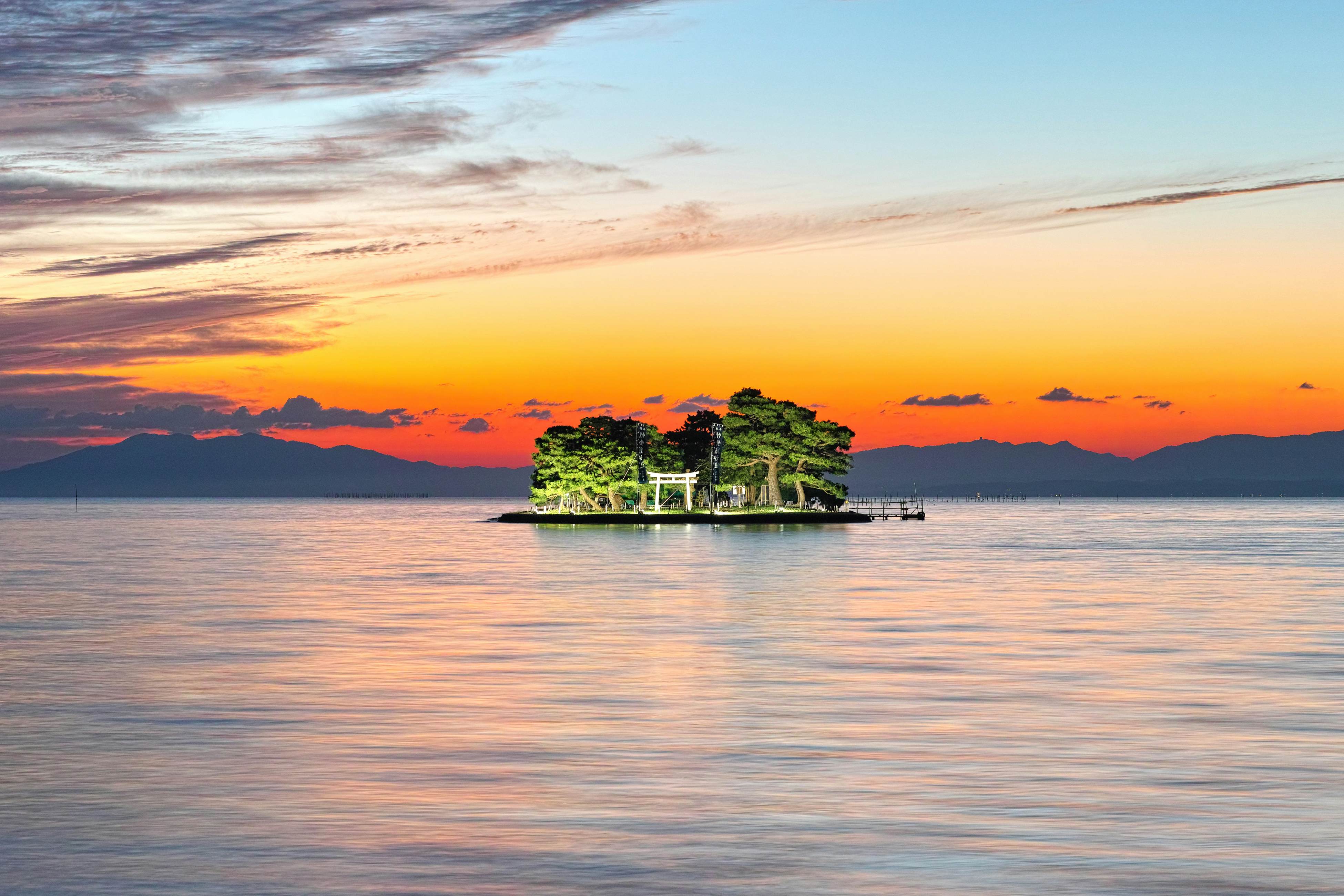 An island with a shrine glows as the sunset streaks the sky with yellows and oranges