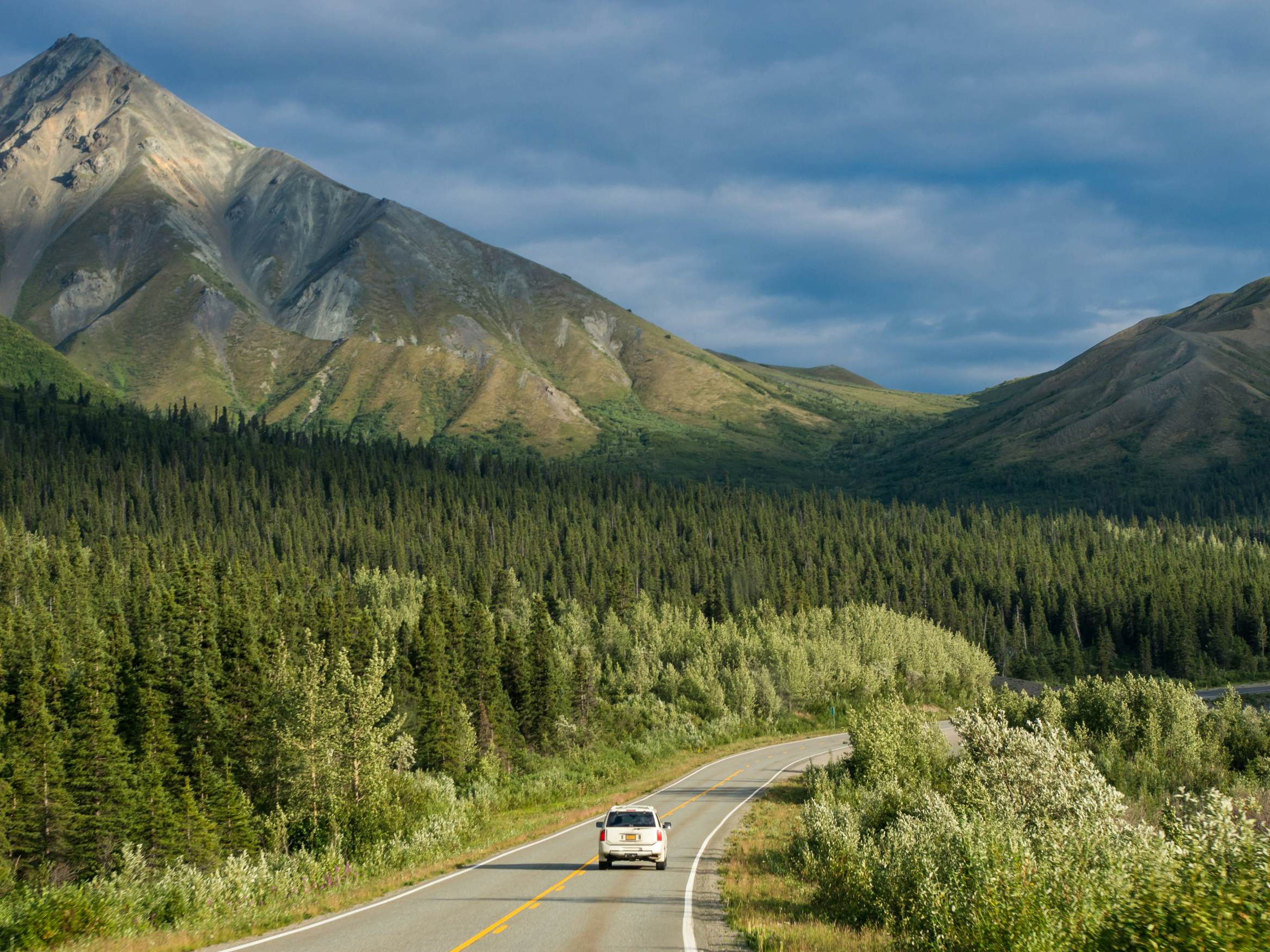 A lone car on a two-way road drives through a grove of pine forests. Huge hills loom in the distance, under a sky filled with dark clouds.