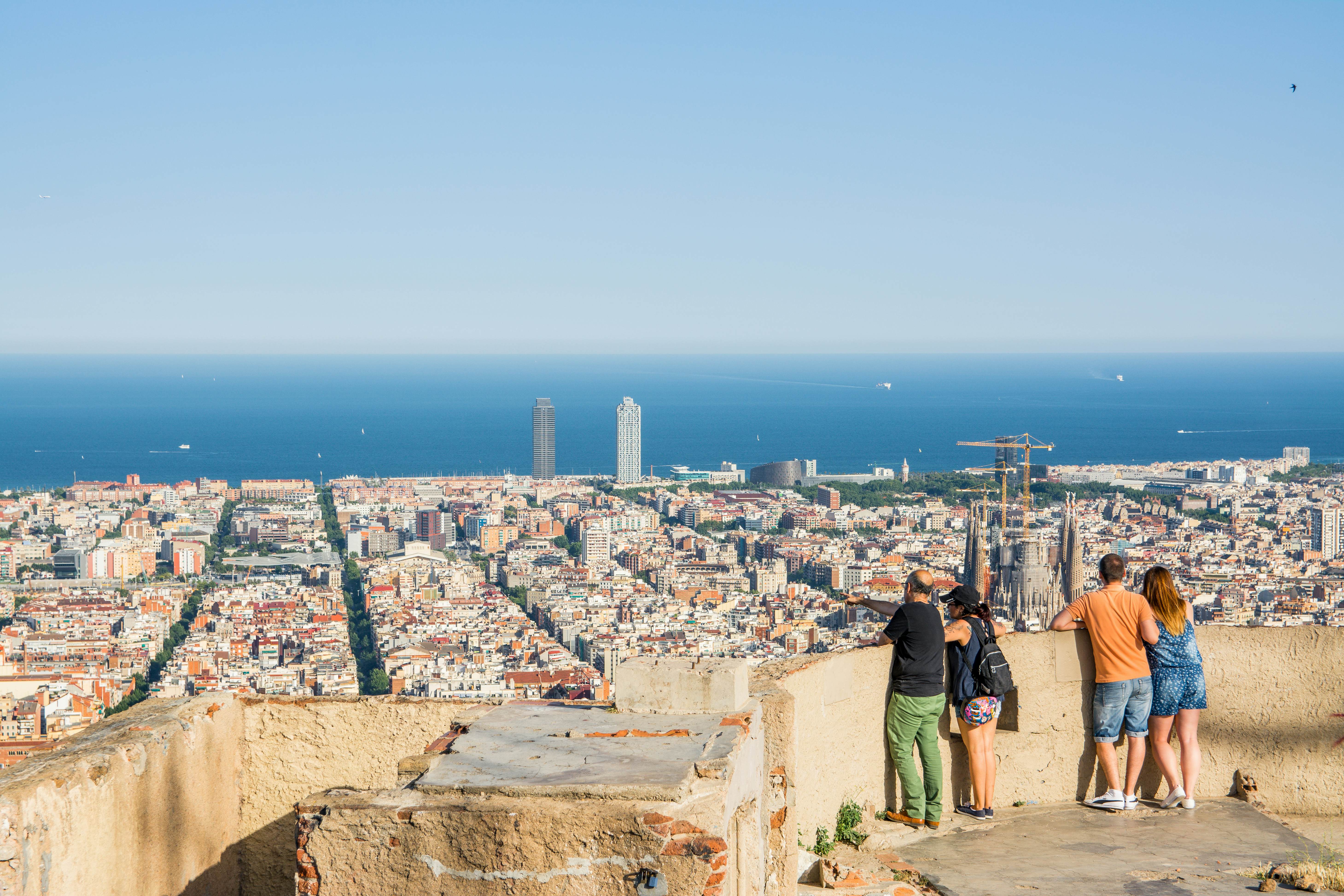 Tourists stand at a viewpoint looking out over the city of Barcelona and the sea.