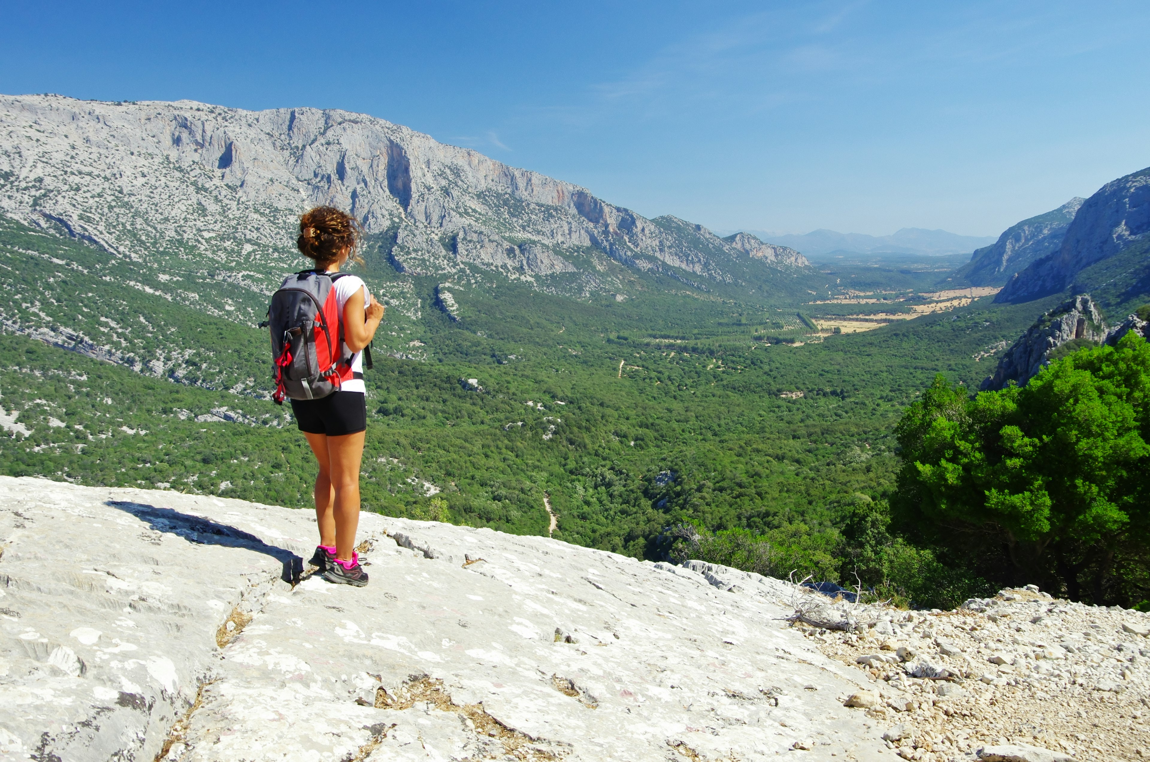 A female hiker wit backpack is seem from behind as she stands on a rock and looks out a a green valley between two ridges of limestone.