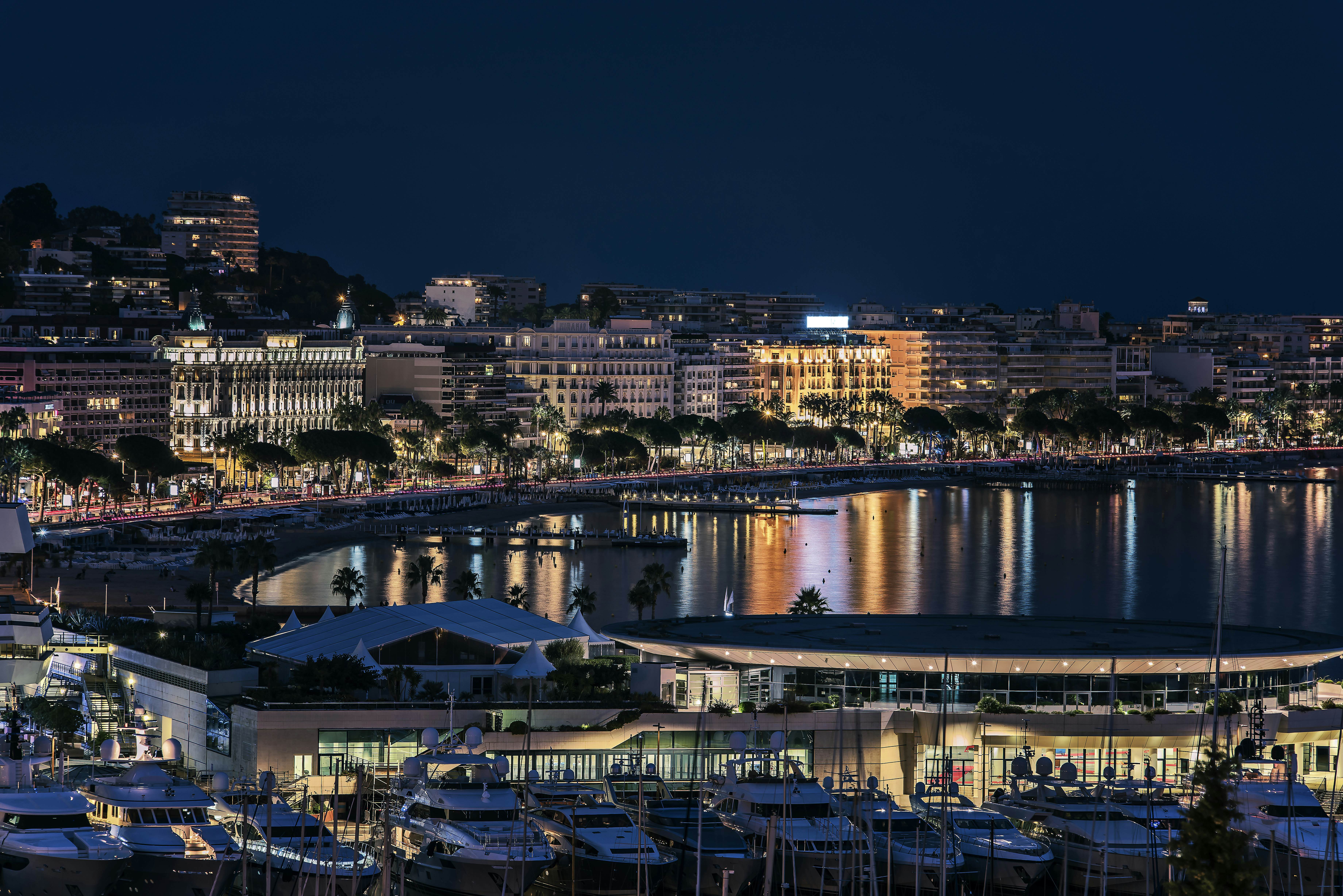 A wide view of a port city by night. Hotels and apartment buildings line the waterfront, their lights reflected in the still water opposite. Boats moored at a pier are visible in the front of the scene.