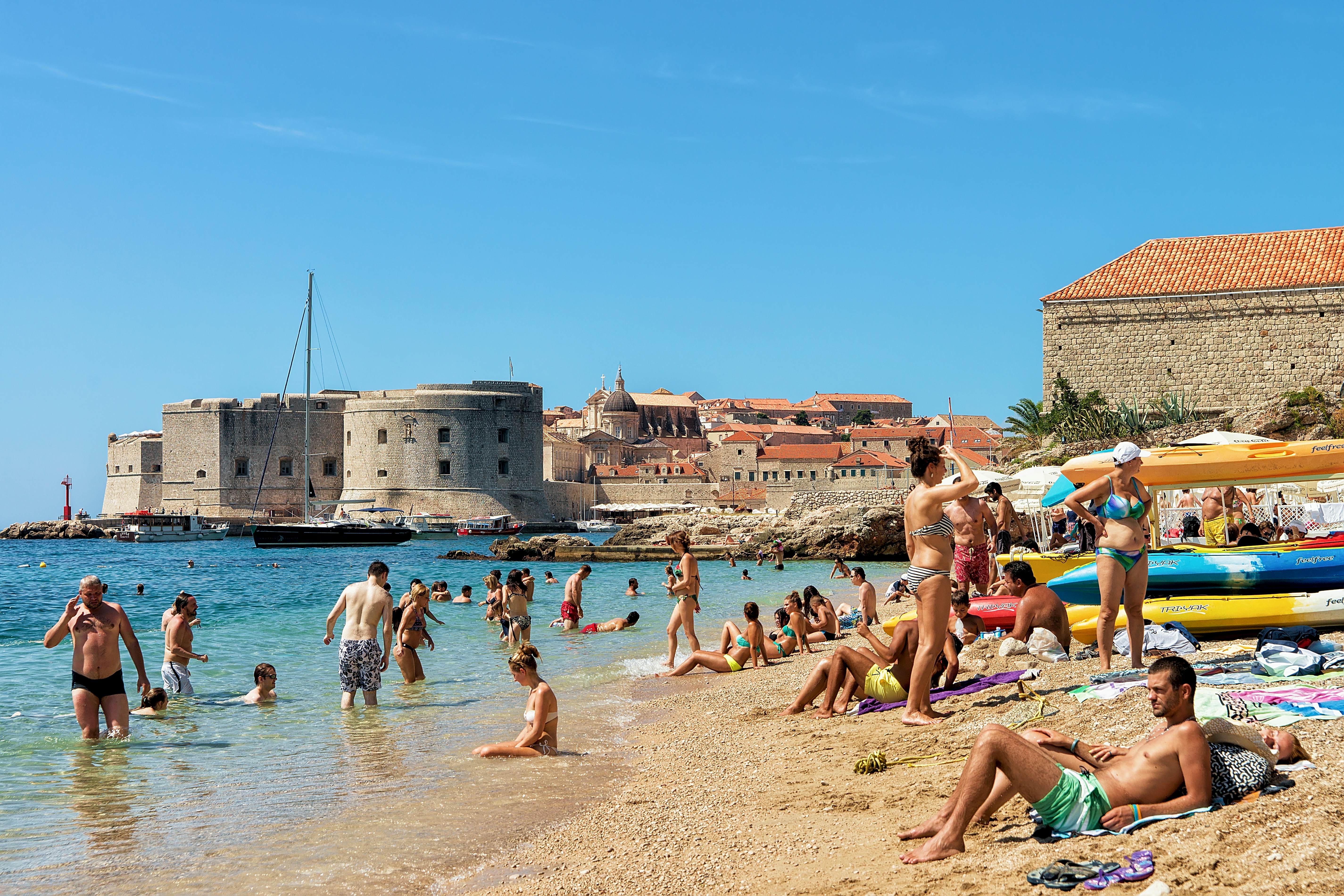 People lie on the sand and wade into the water at a beach by a historic town. A fortress is visible across the water.