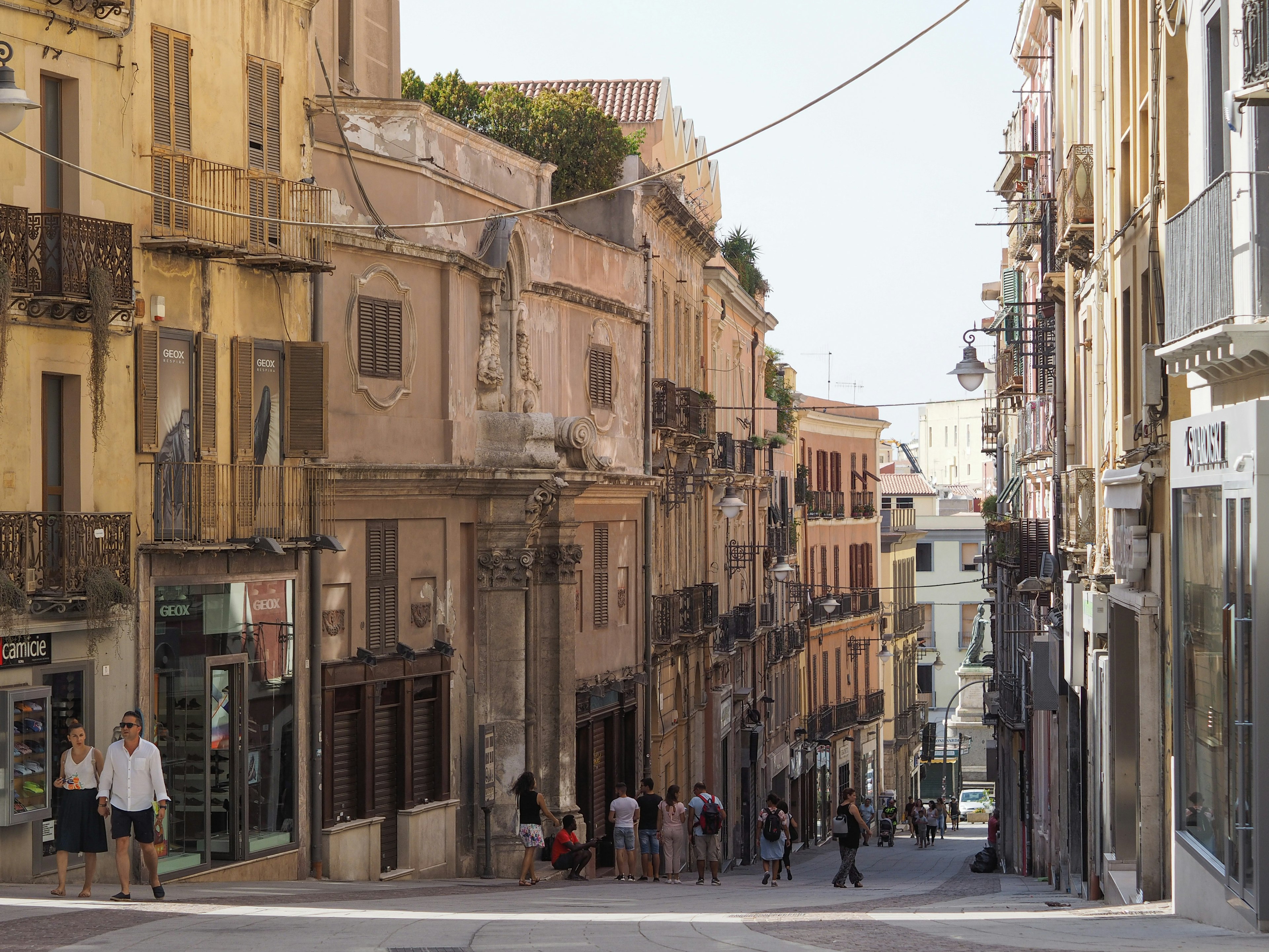 People walk down a pedestrianized street by historic buildings with shops and balconies.