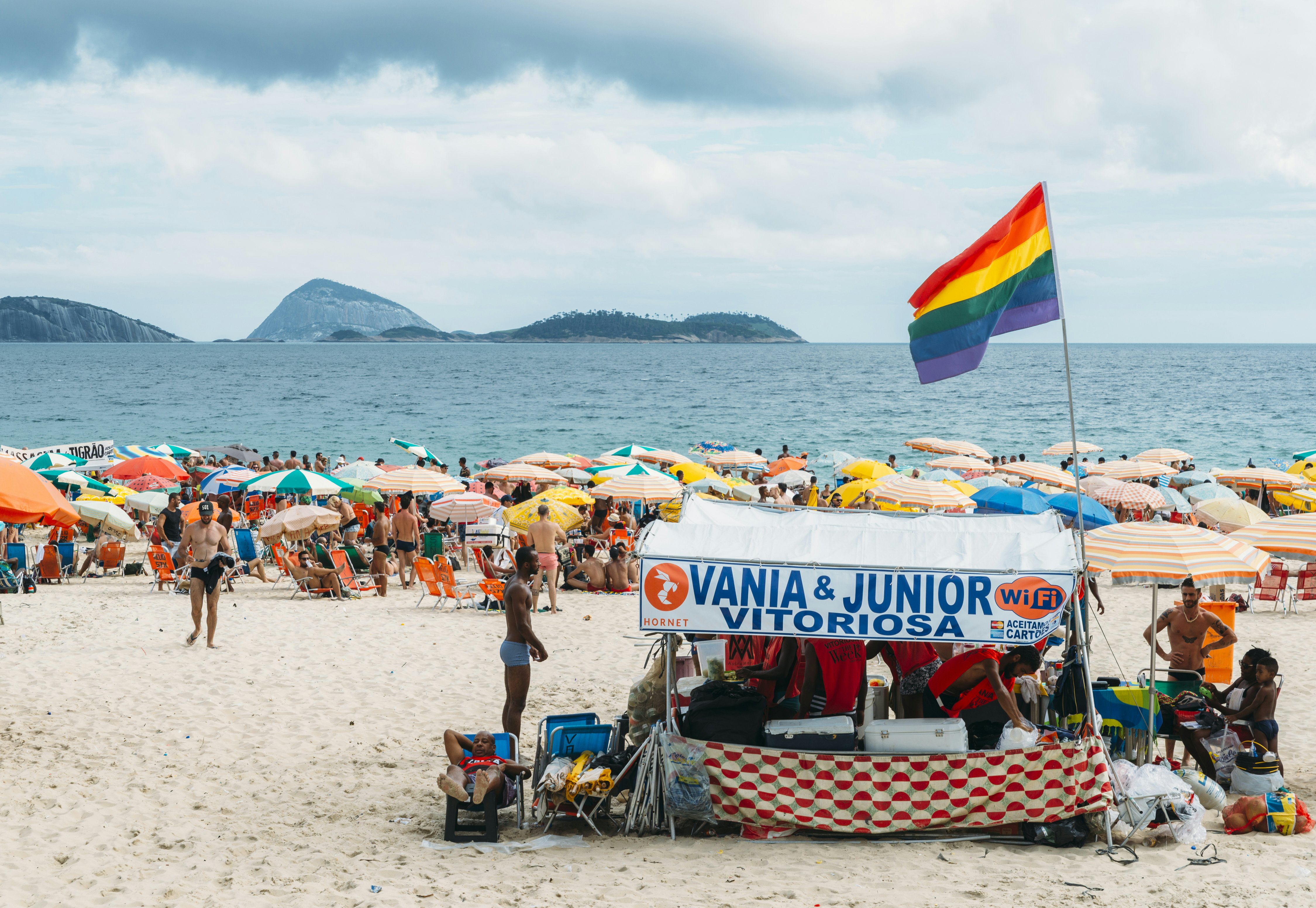 A vendor flying a rainbow flag is pictured on a busy city beach. Hundreds of beach umbrellas and sunbathers can be seen closer to the water.