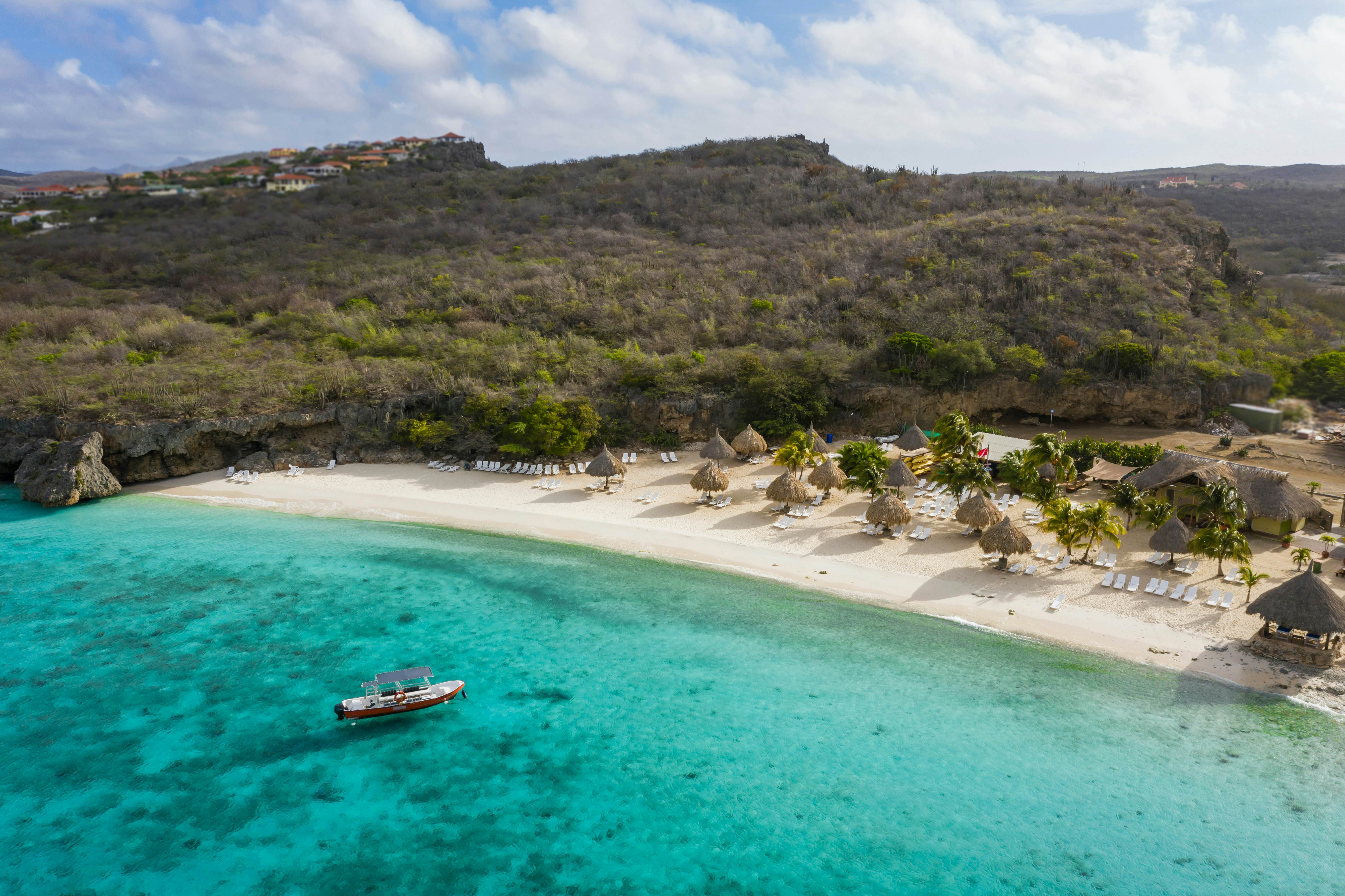 A boat moored in the turquoise ocean near a large beach with sunshades and loungers