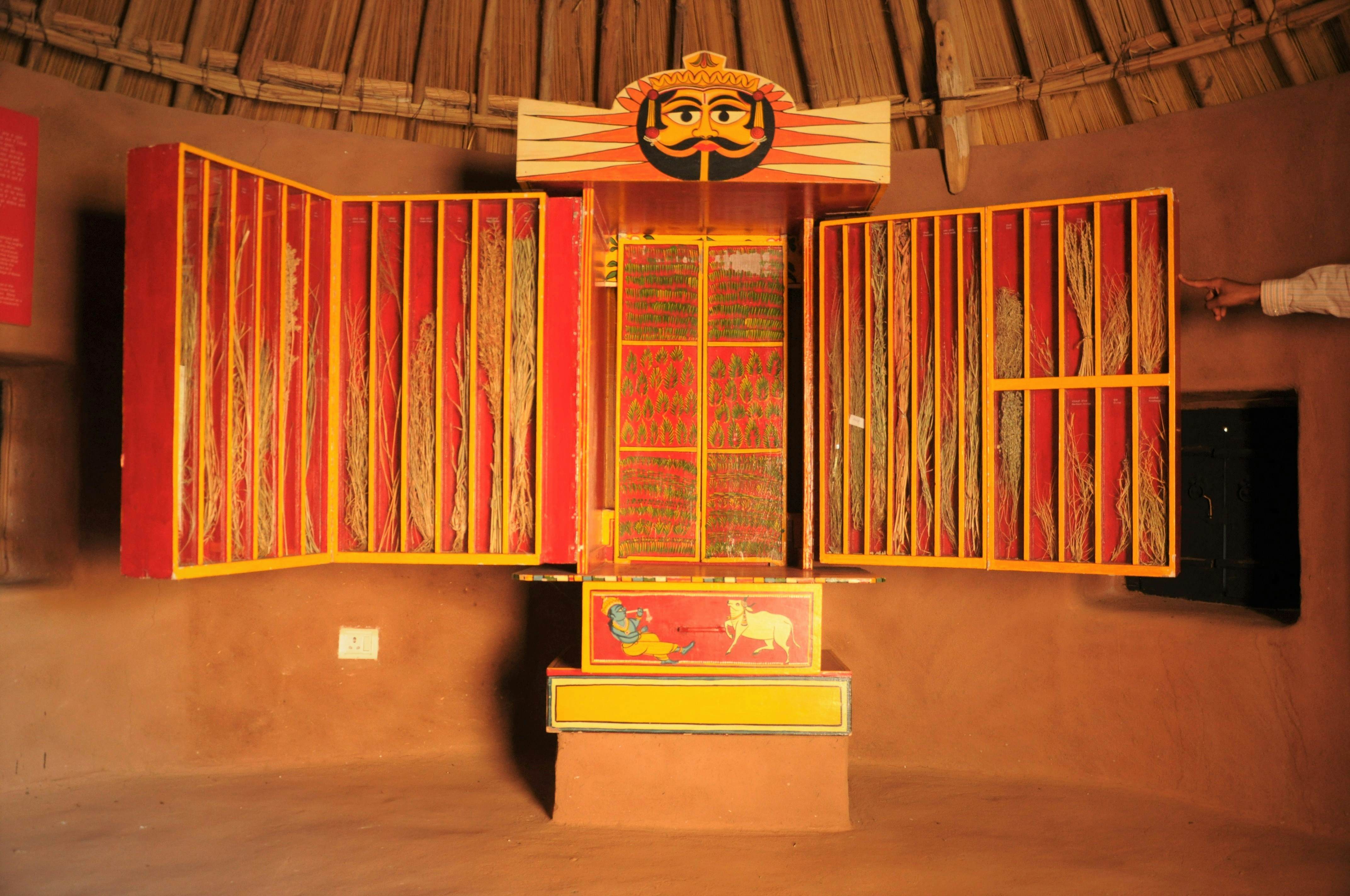 A painted display case at a museum contains brooms of various sizes. A panel with a painted head is mounted above the case.