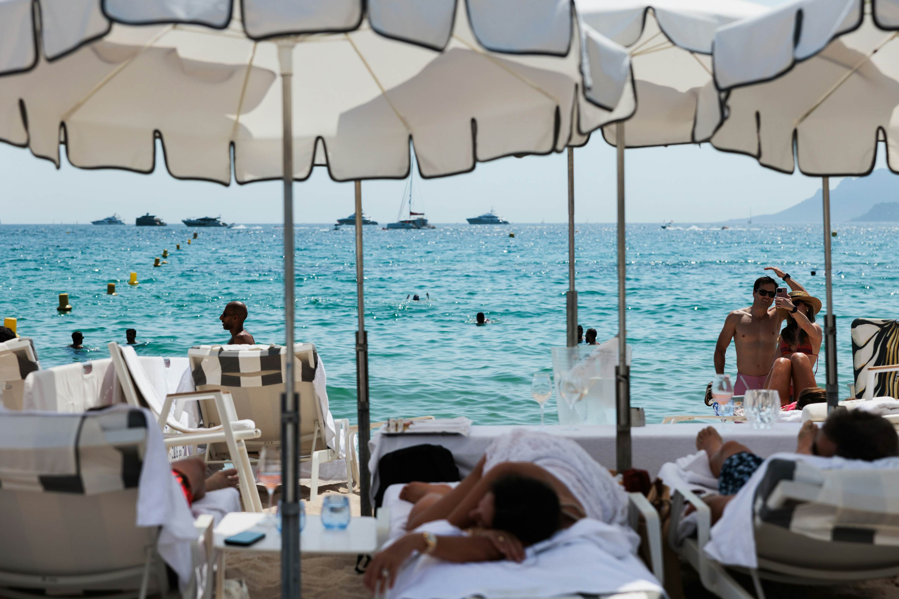 A view of people lounging on chairs under umbrellas on a beach. People swim in the blue water just offshore, and yachts can be spotted moored in the distance.