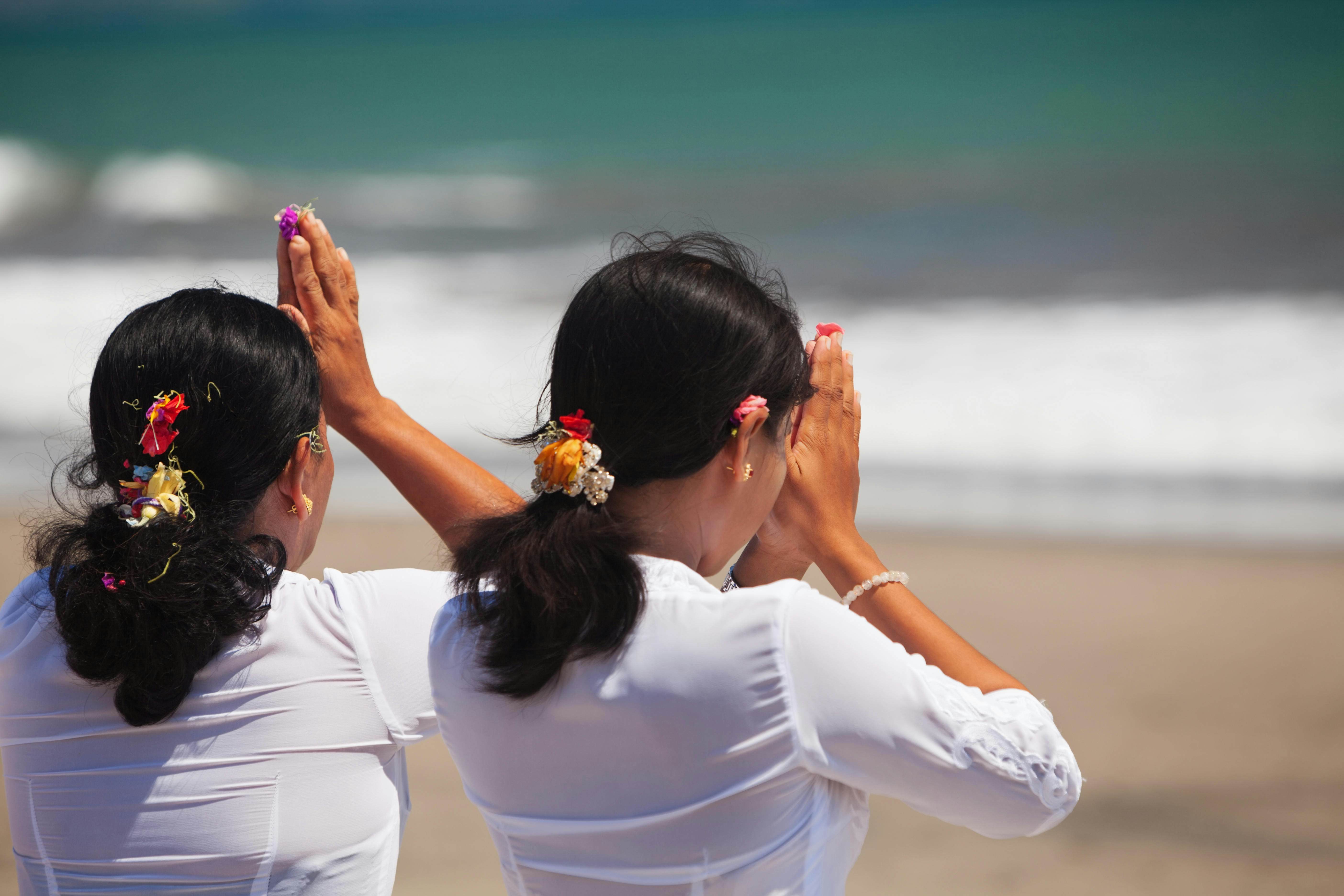 Two women with flowers in their hair kneel on a beach in prayer, their hands by their heads.