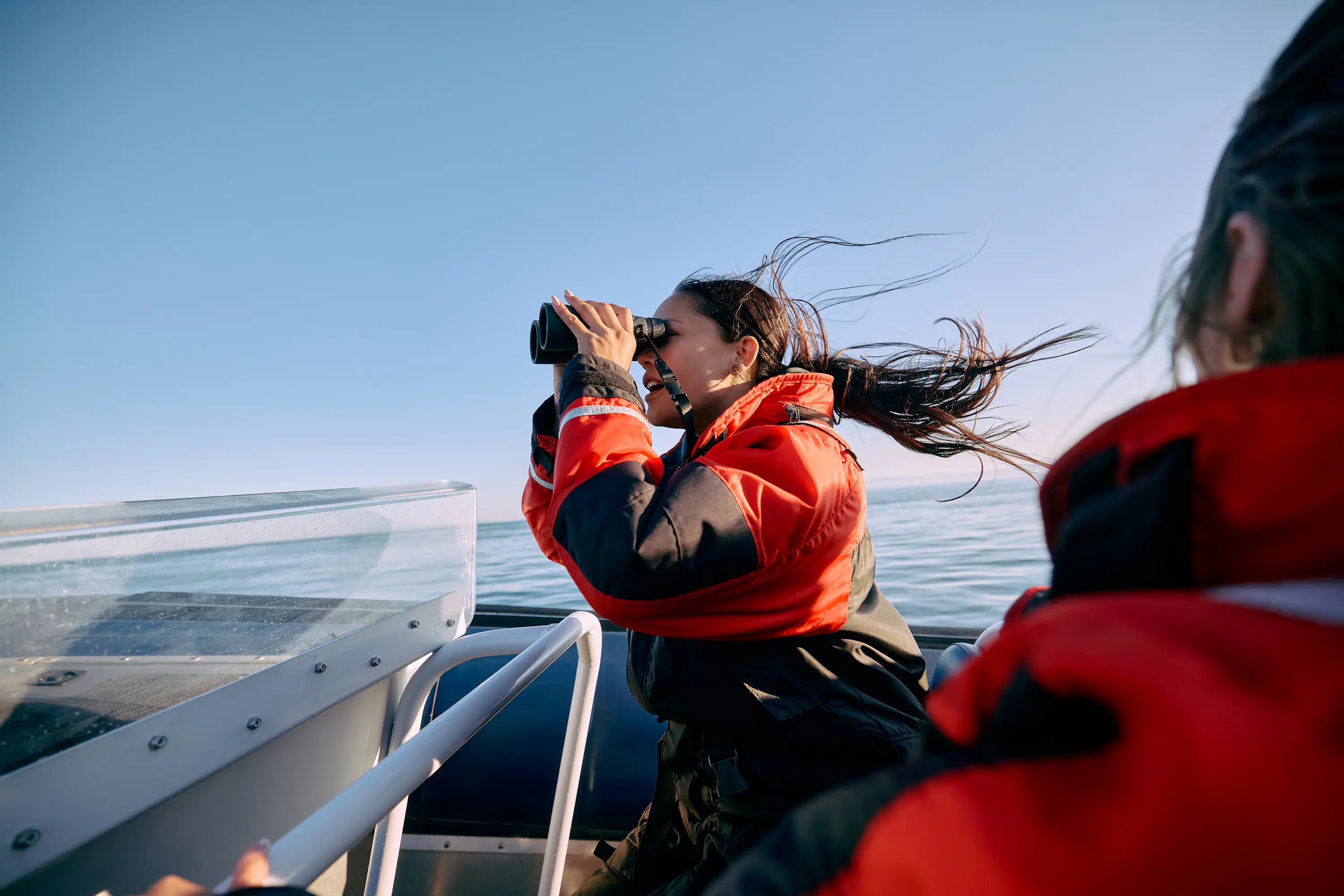 A woman in an orange jacket on a boat uses binoculars to look for whales off the coast of British Columbia