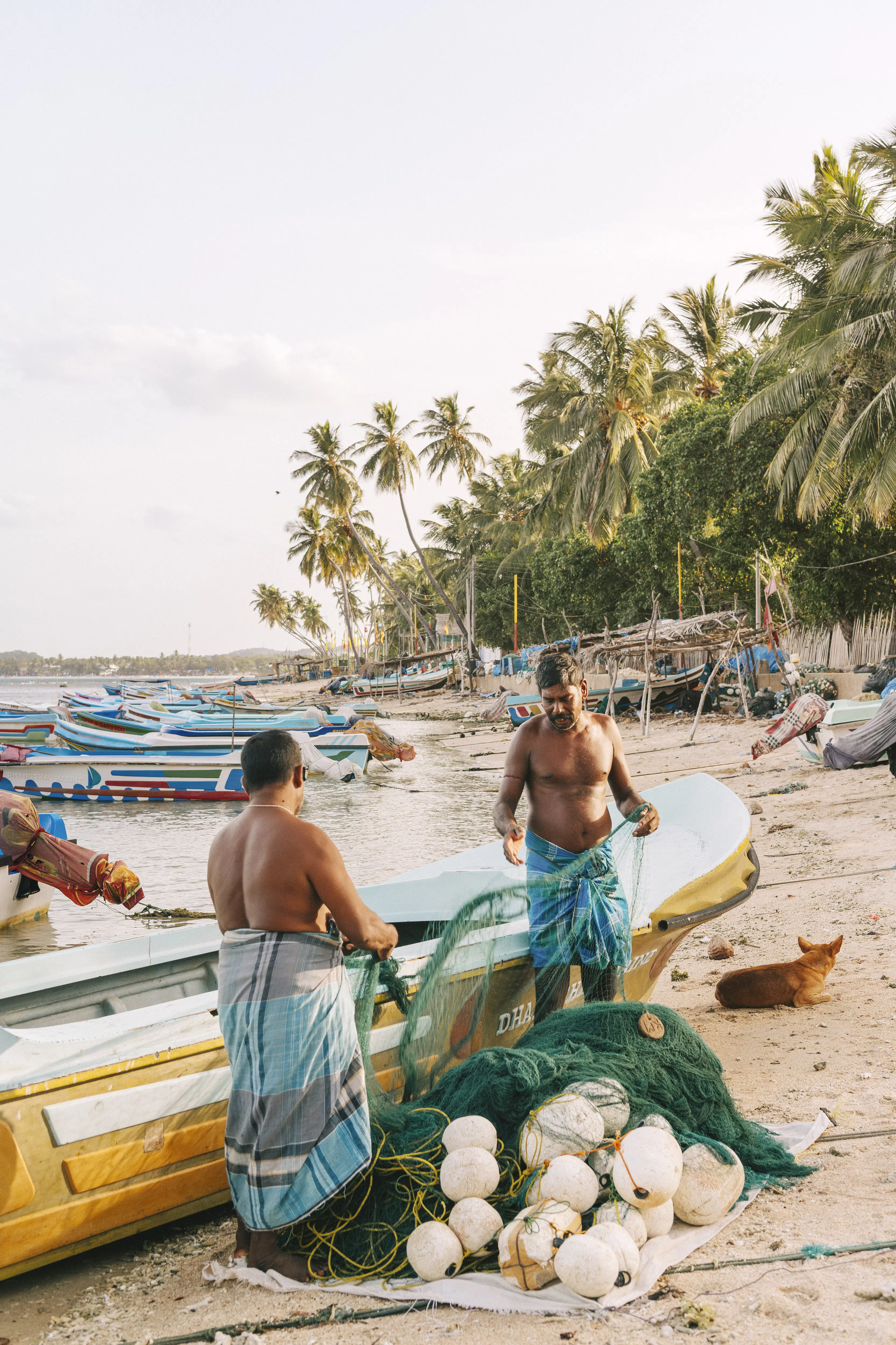 Two men inspect fishing nets by a boat on the sand.