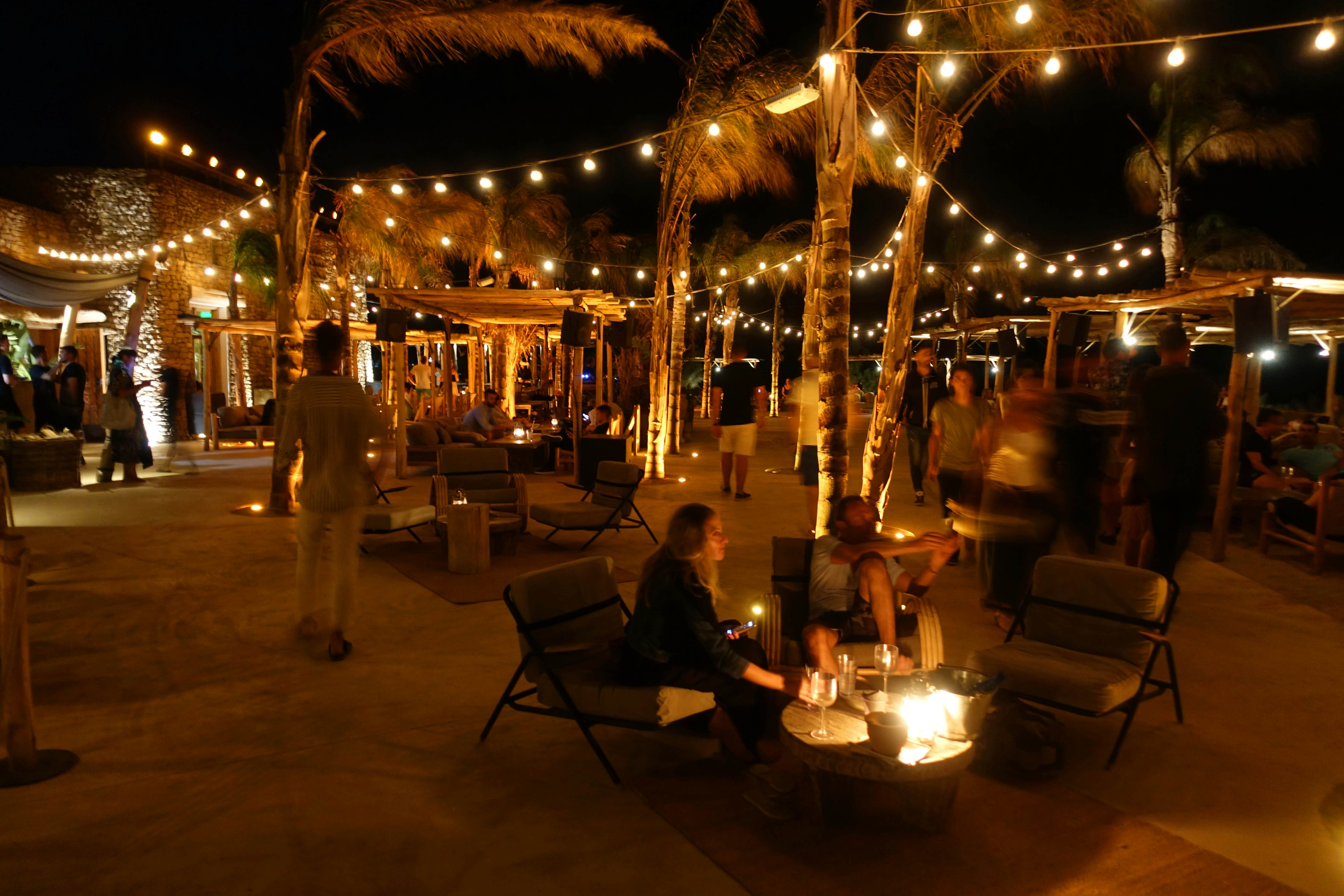 A beach bar at night, with hanging lights and small groups seated around low tables, in Mykonos, Greece