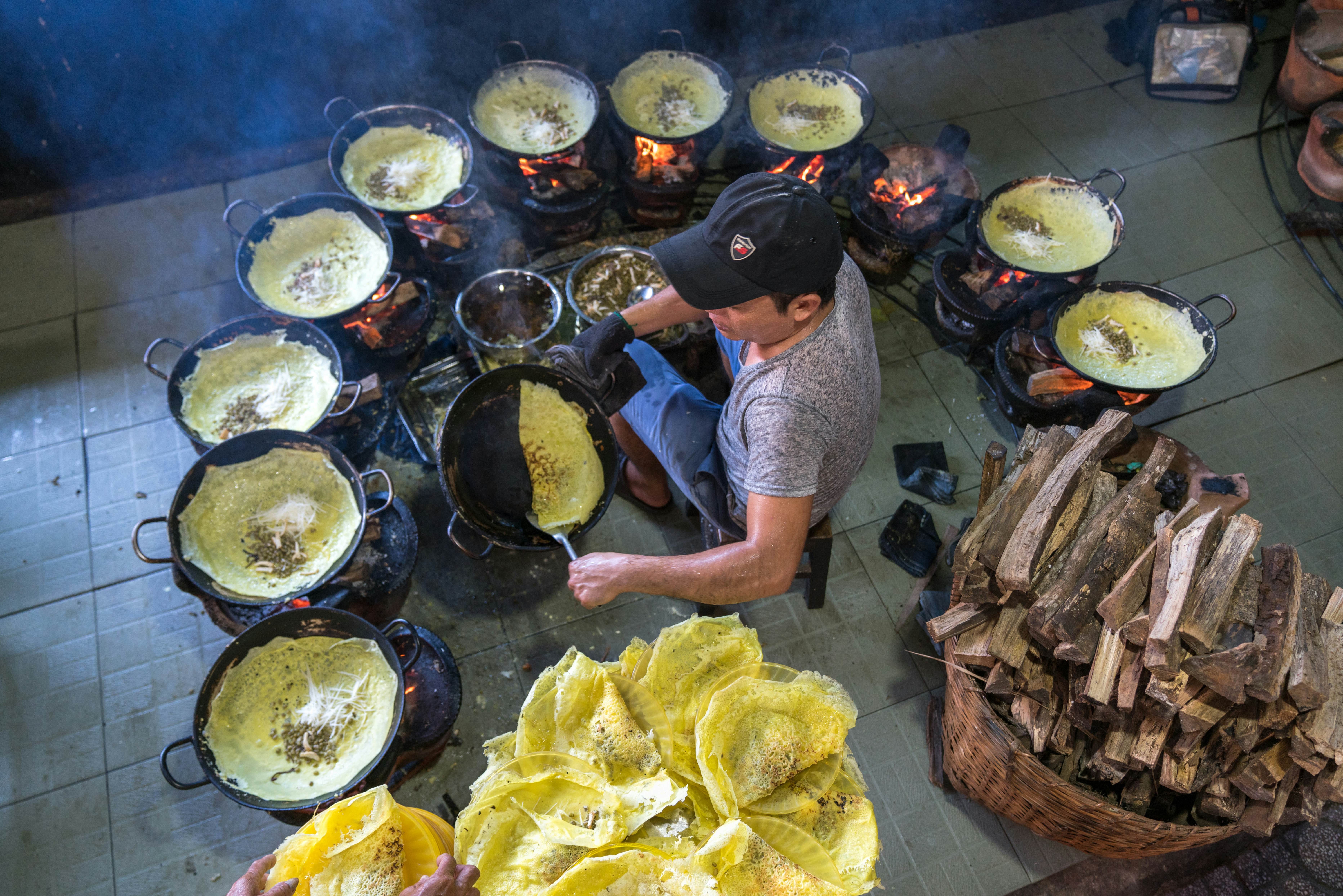 An overhead shot of man sitting while cooking banh xeo (Vietnamese sizzling cake) on a sidewalk. He is cooking with several skillets at once. Wood for the fire is seen behind him.