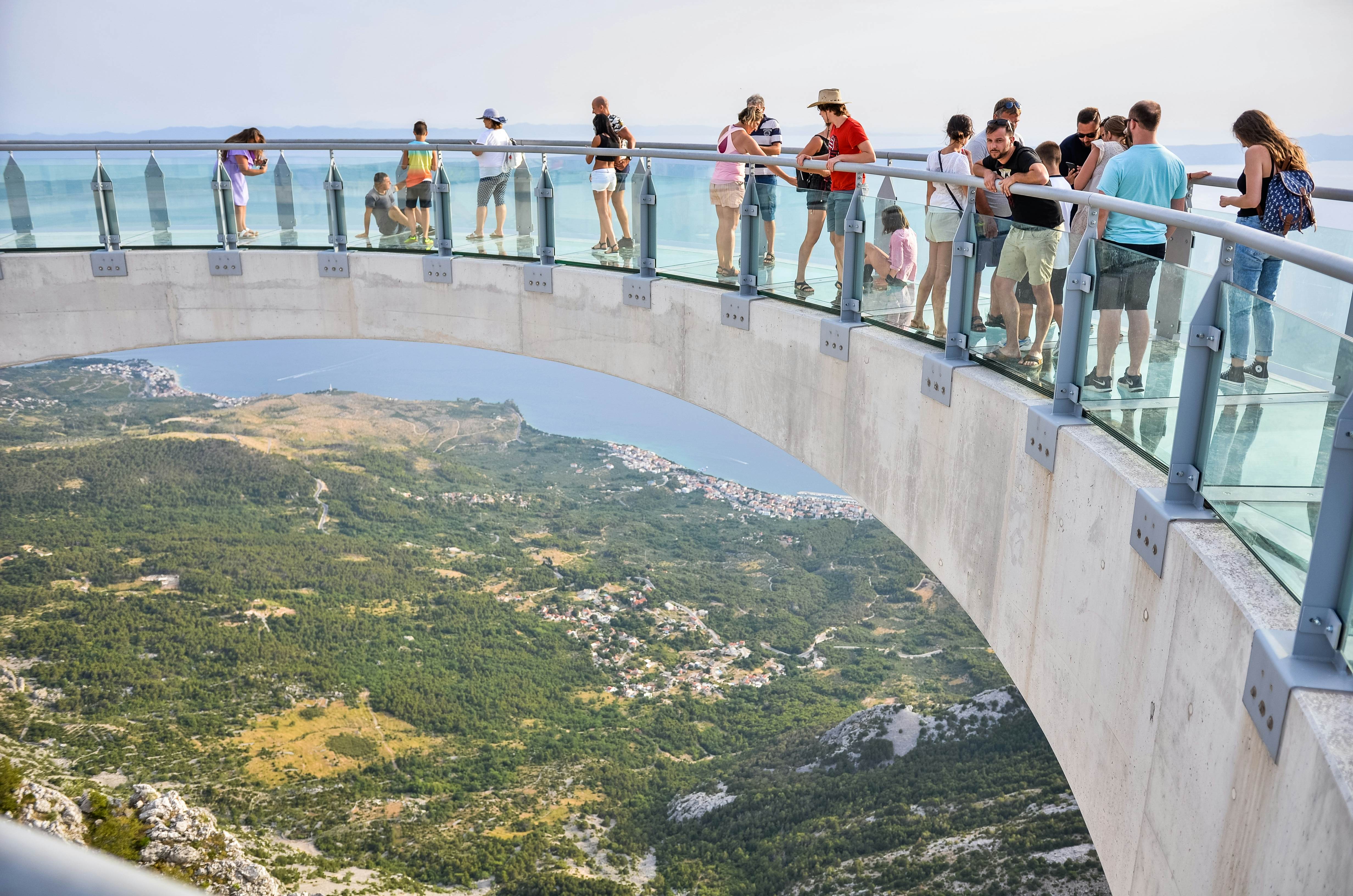 People stand on a curved walkway that juts out over a cliff, with a view of the valley and sea below.