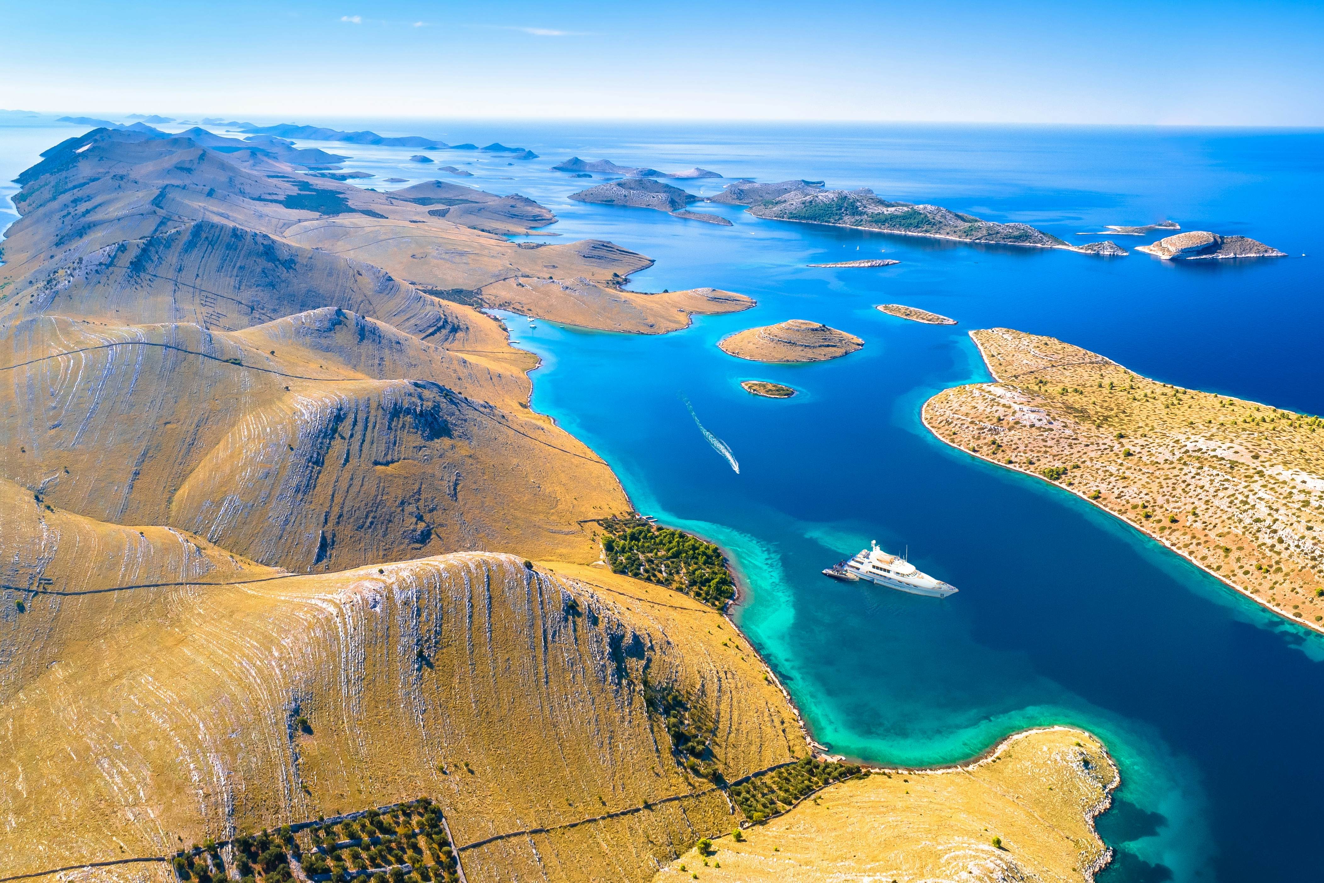 An aerial view of a yacht navigating the waters between hilly islands with scrubby vegetation.