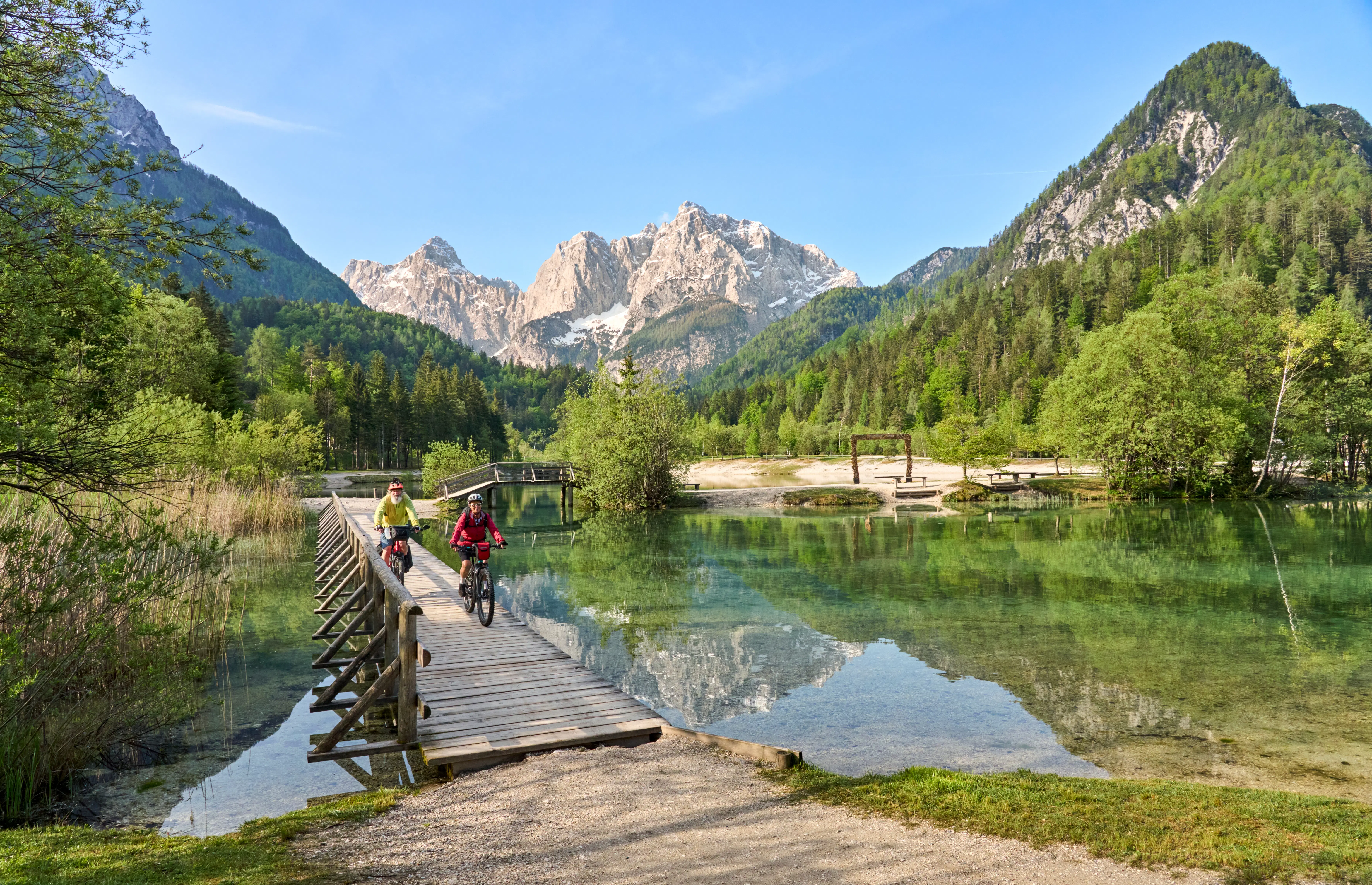 Two cyclists pedal along a boardwalk over lakes in a mountainous region.
