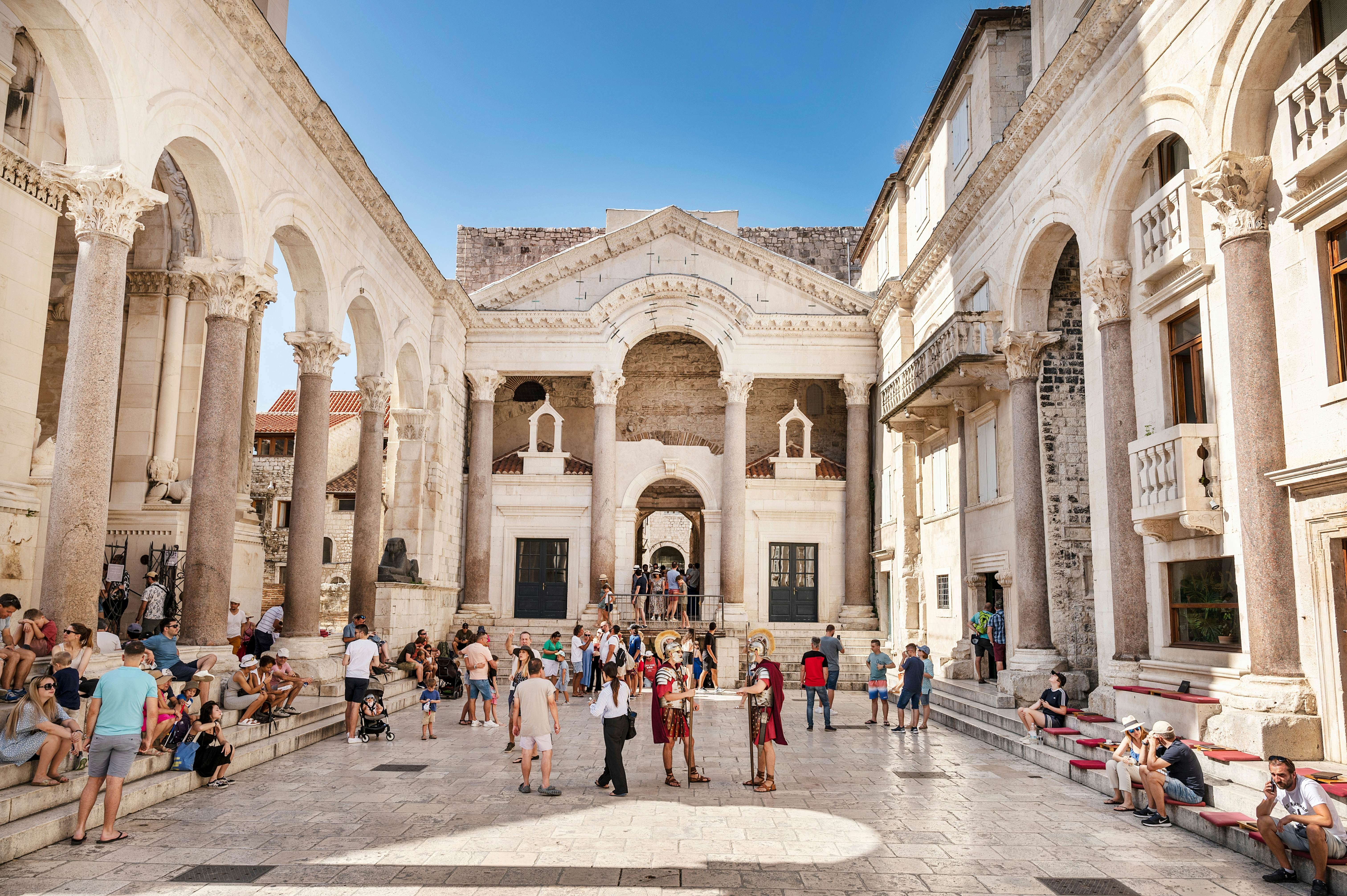 People gather in a courtyard of an ancient building with numerous columns, balconies and other architectural details.