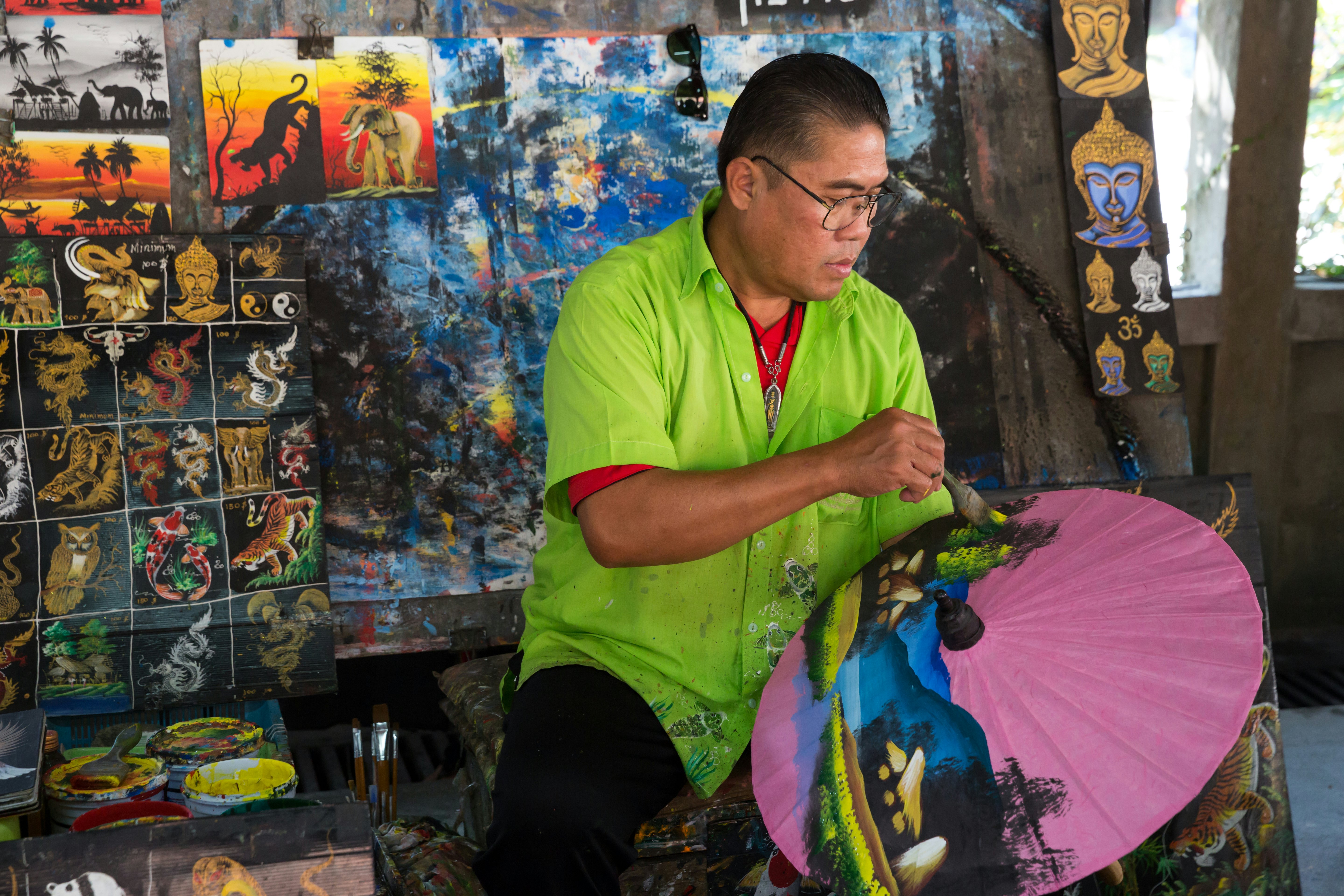 An artisan in a bright green shirt carefully paints an umbrella in a workshop.