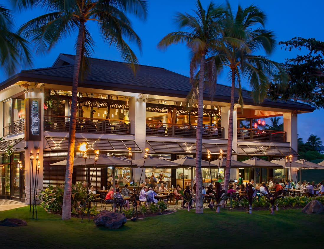 Evening diners at tables under palm trees at the two-story Monkeypod Kitchen at the Ko Olina Center