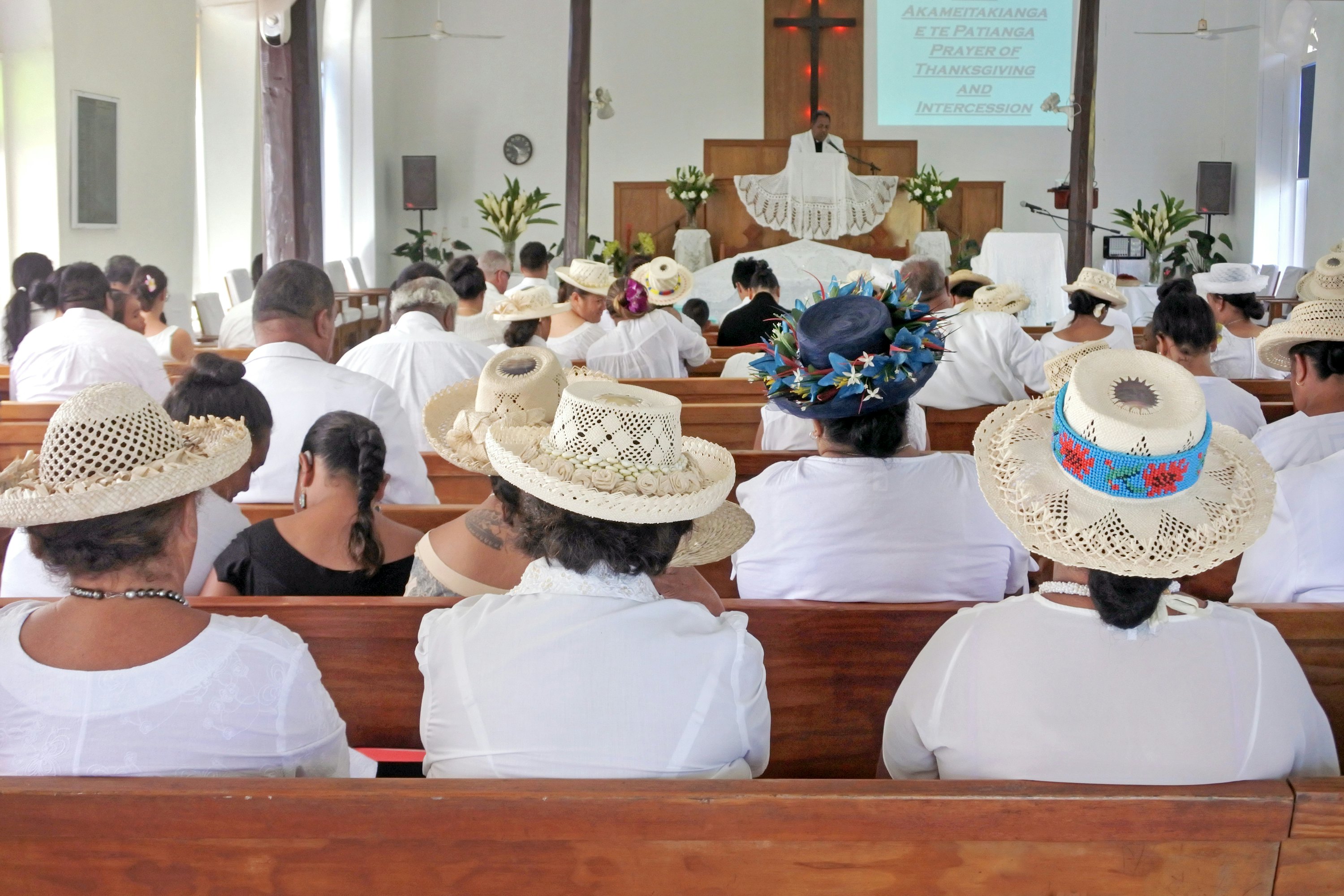 Women wearing white outfits and hats are seen from behind sitting in pews in a church during a Sunday service.