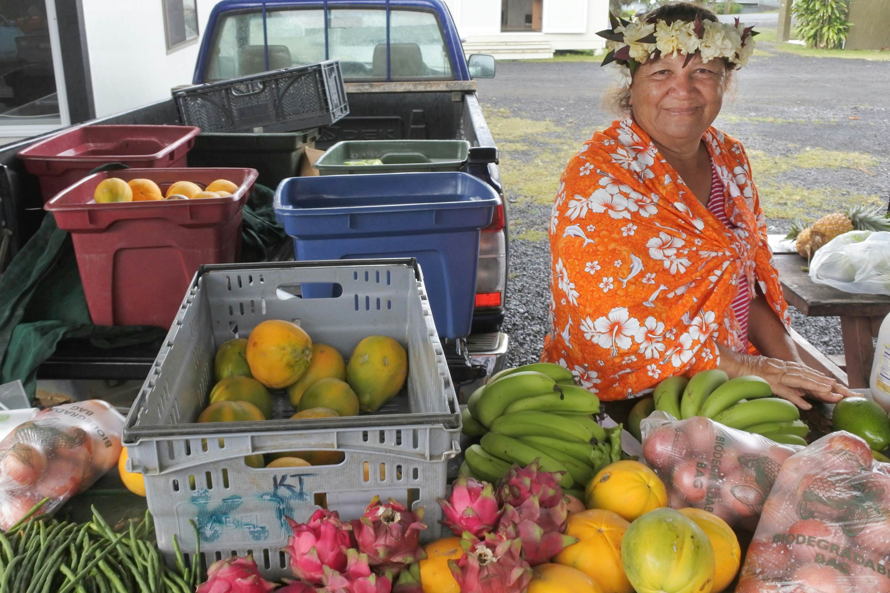 A woman with an orange shawl and flowers in her hair sells fresh fruits at a market in a town.