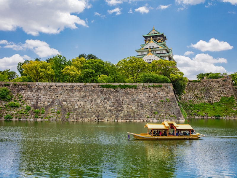 A traditional Japanese structure rises above green trees that are planted above a stone wall. The stone wall abuts a river on which a boat carries tourists.
