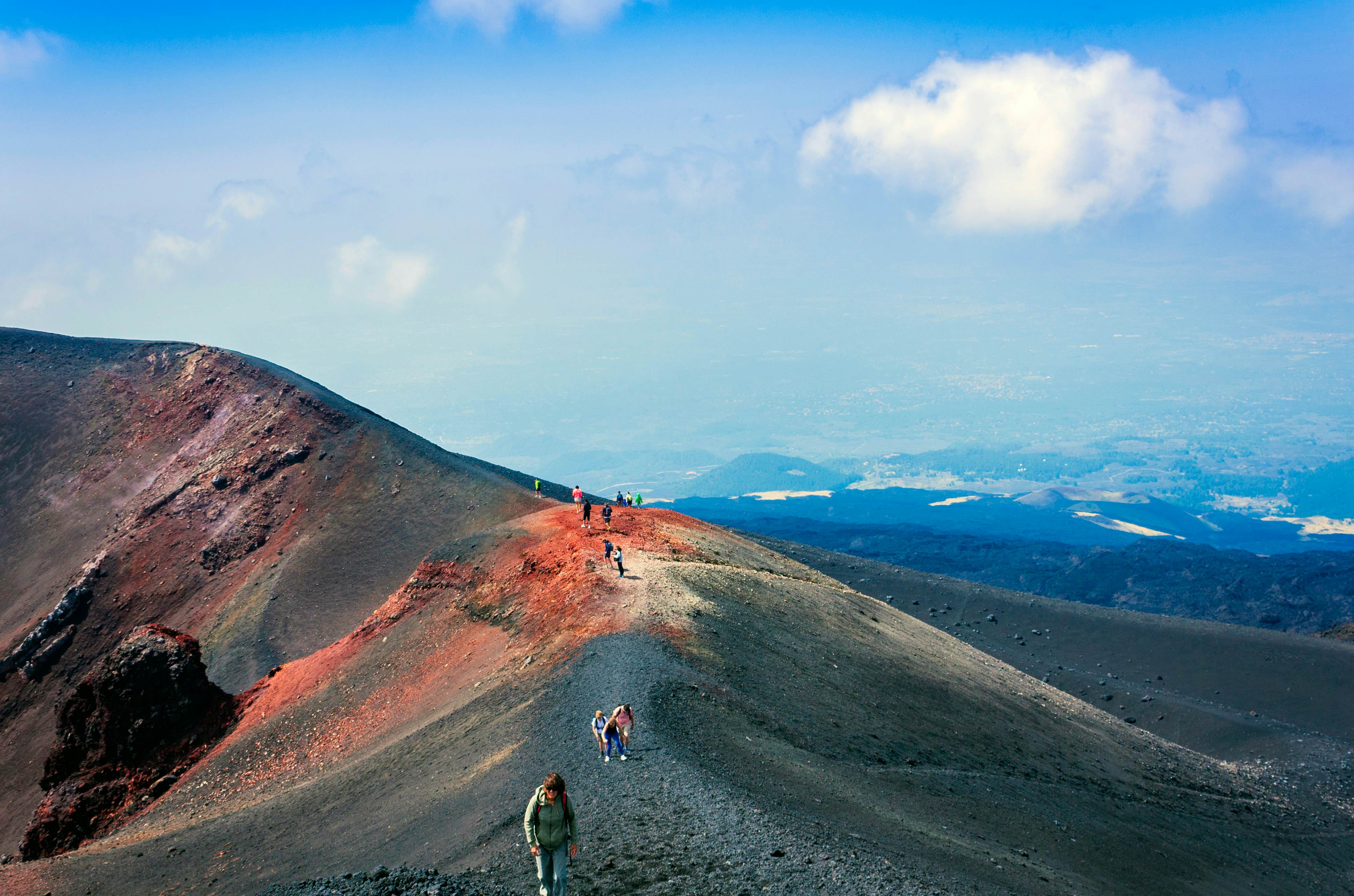 People walk along the ridge of a dusty mountain. Light makes the slopes appear red; hills and fields are visible far below, in the distance.