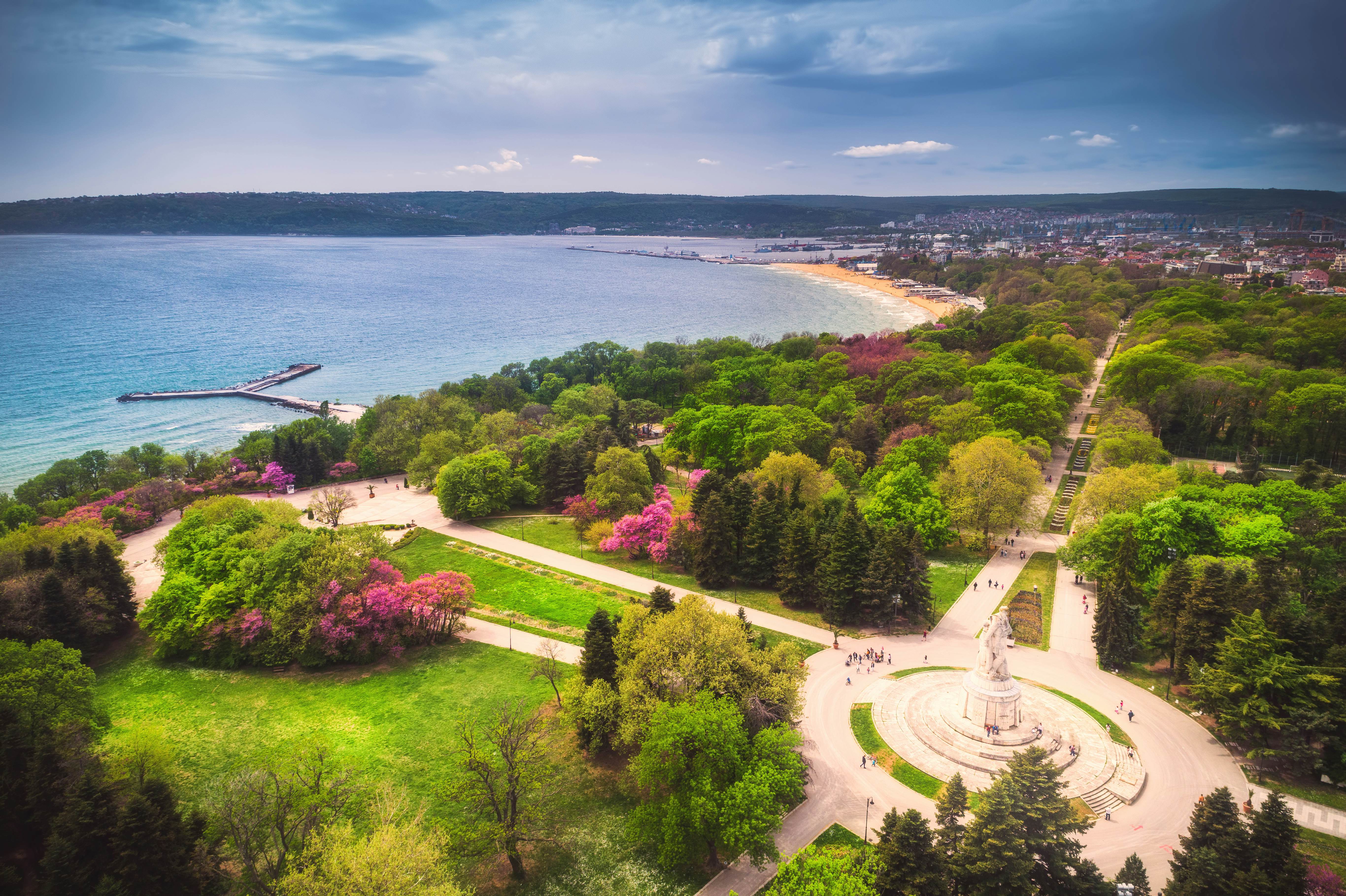 A huge park with trees, pathways, statues and flower beds runs alongside the sea, with a sandy beach in the distance