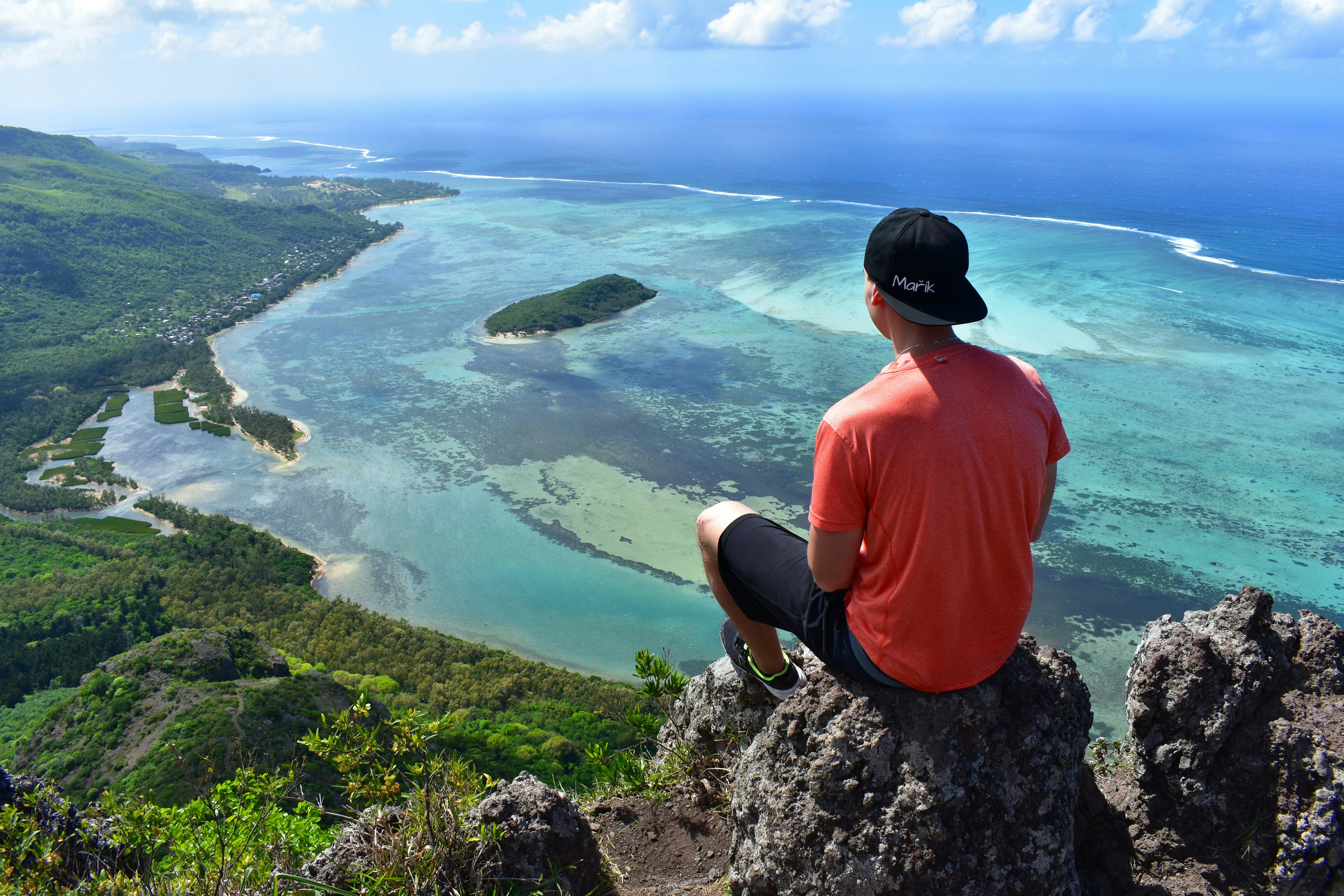 A male hiker is seen from behind, sitting on a rock at the top of a mountain, looking out at the blue sea and lush landscape below.