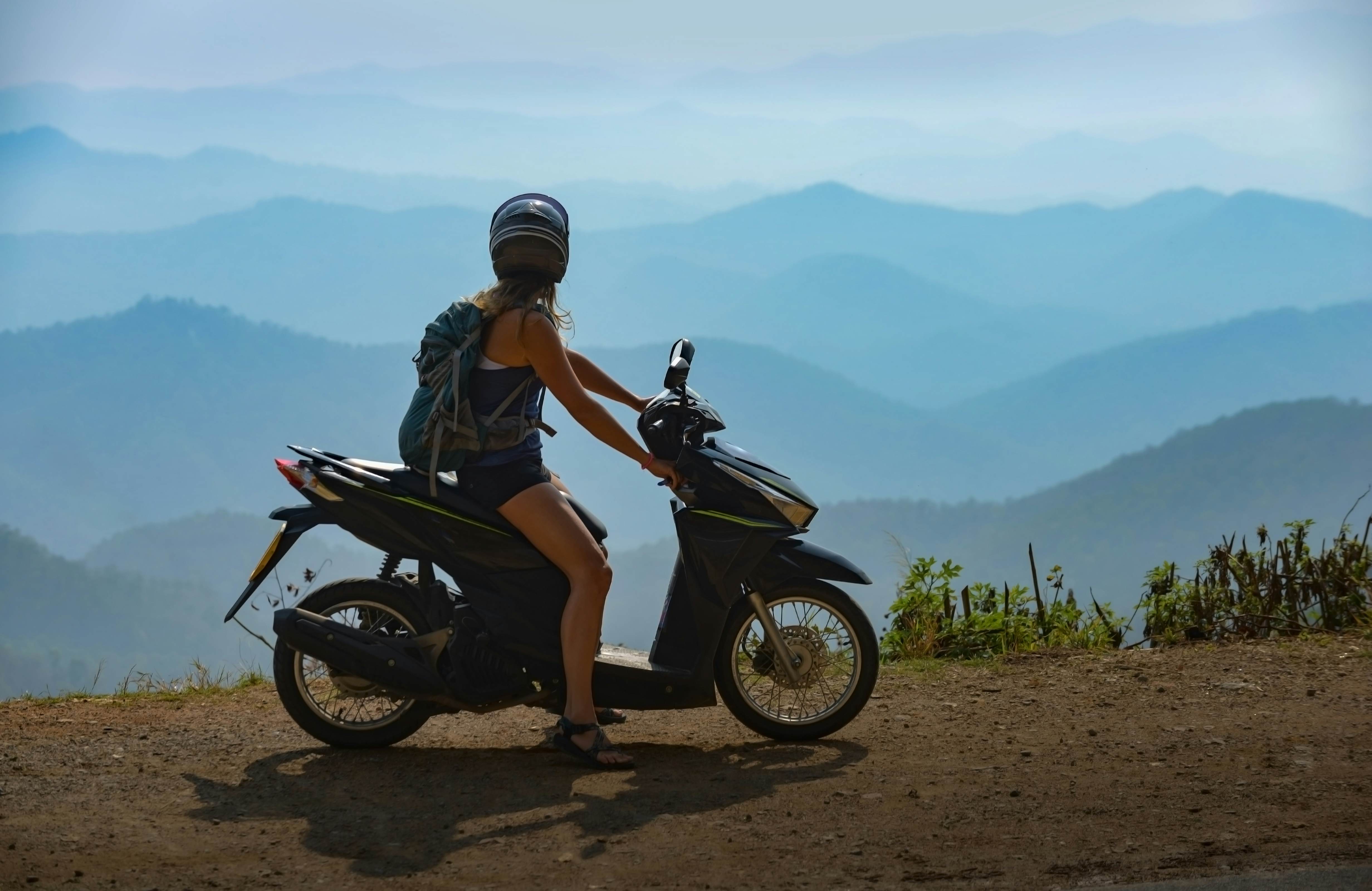 A female on a motorbike stops to admire a view of misty blue mountains on a road on a mountain pass.