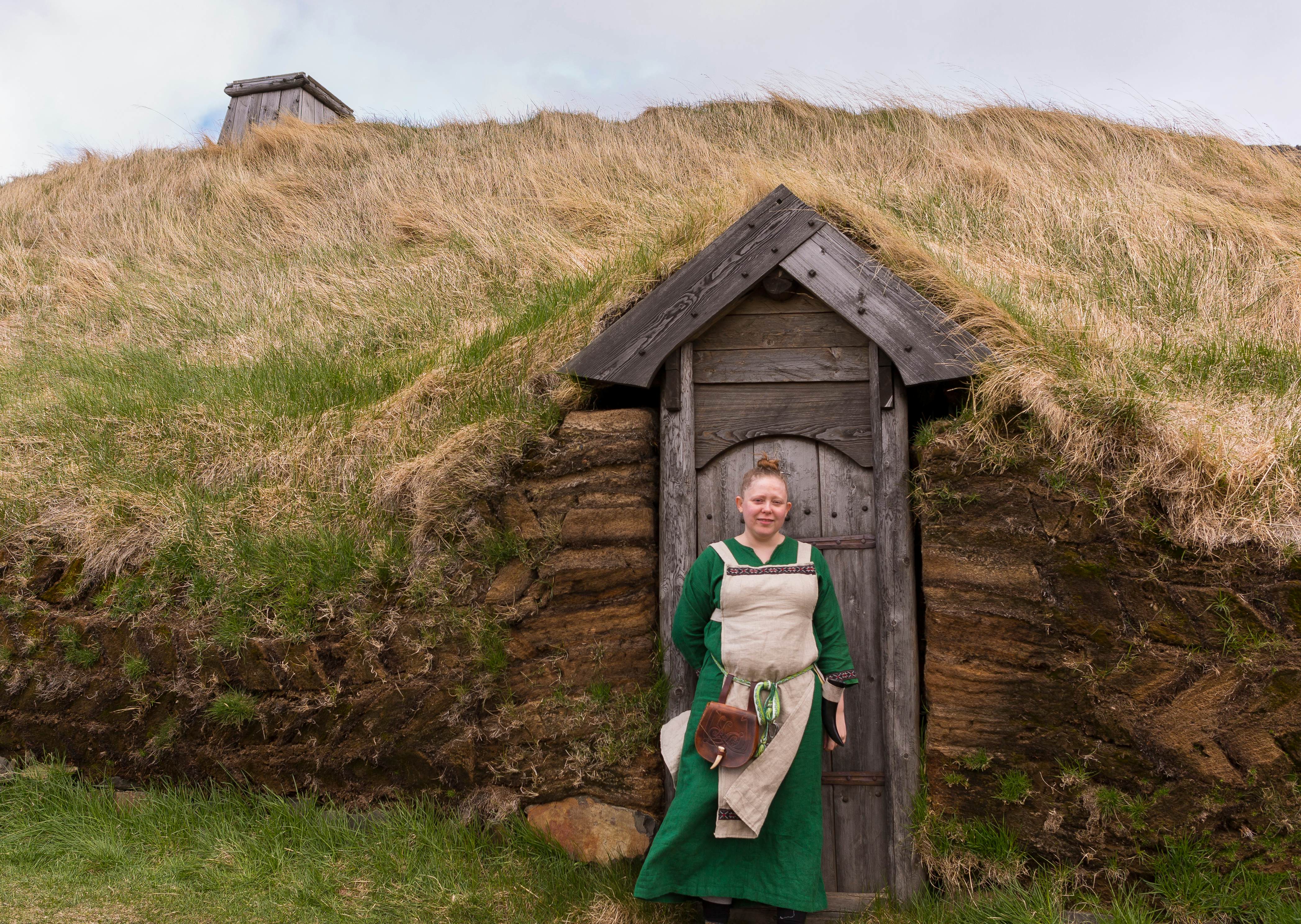 A woman in a green dress and period costume stands in front of a wooden door to a structure covered whose roof is earth and grass.