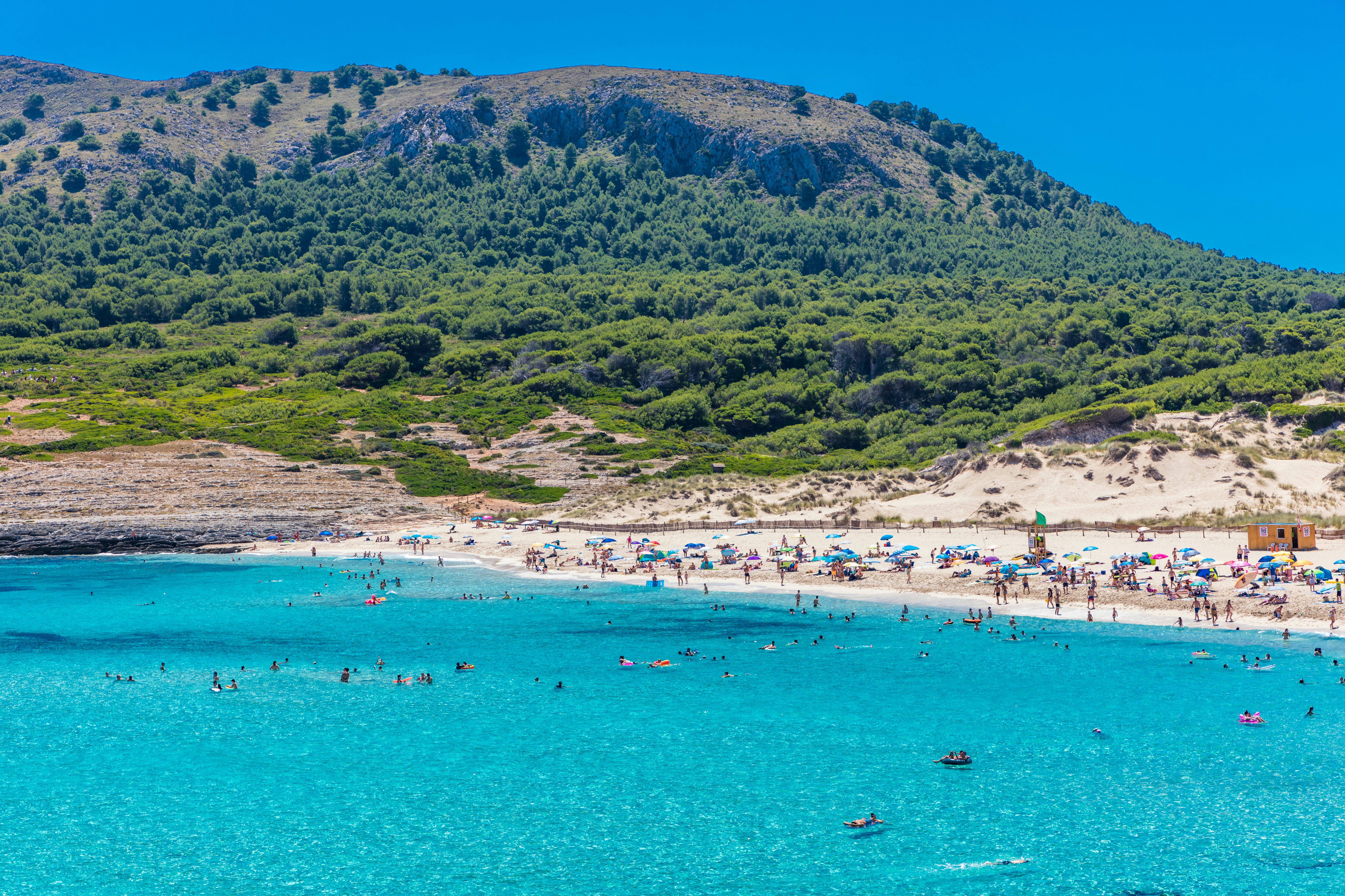 People swim in brilliant blue waters surrounded by a sandy beach and forested hills