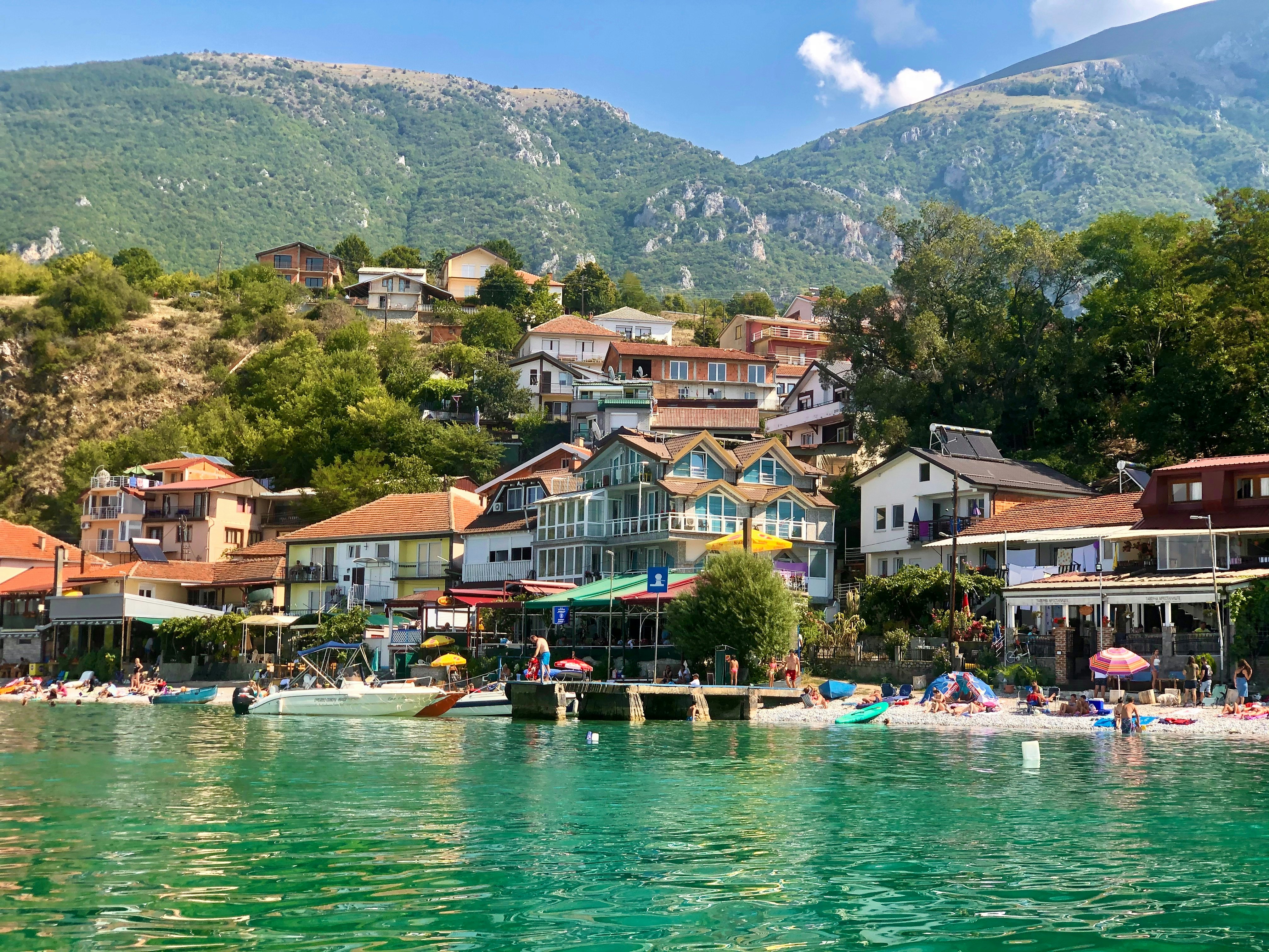 A small lakeside village backed by mountains with people relaxing on the sandy beach