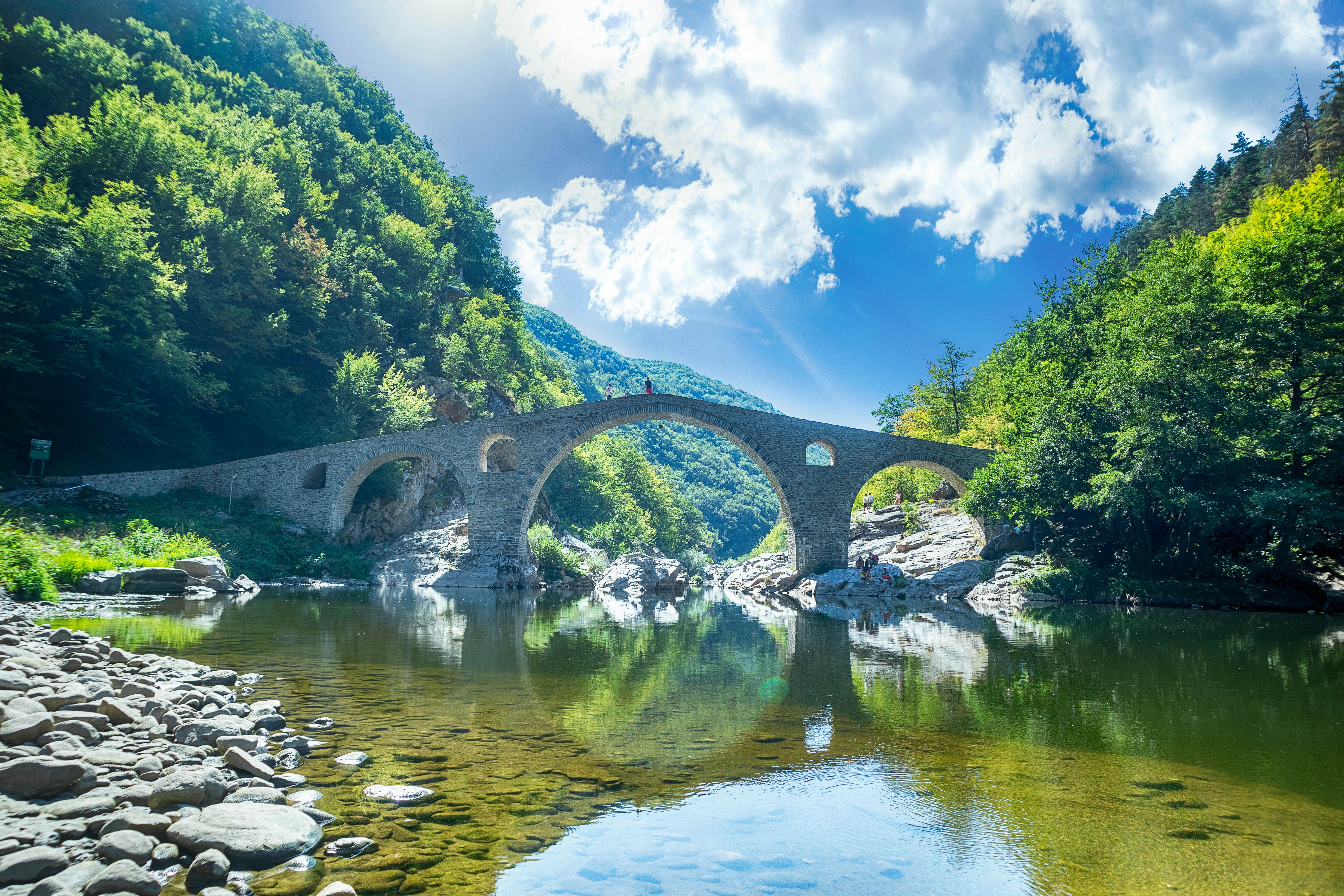 An ancient stone bridge crosses a glacial river with mountains in the distance