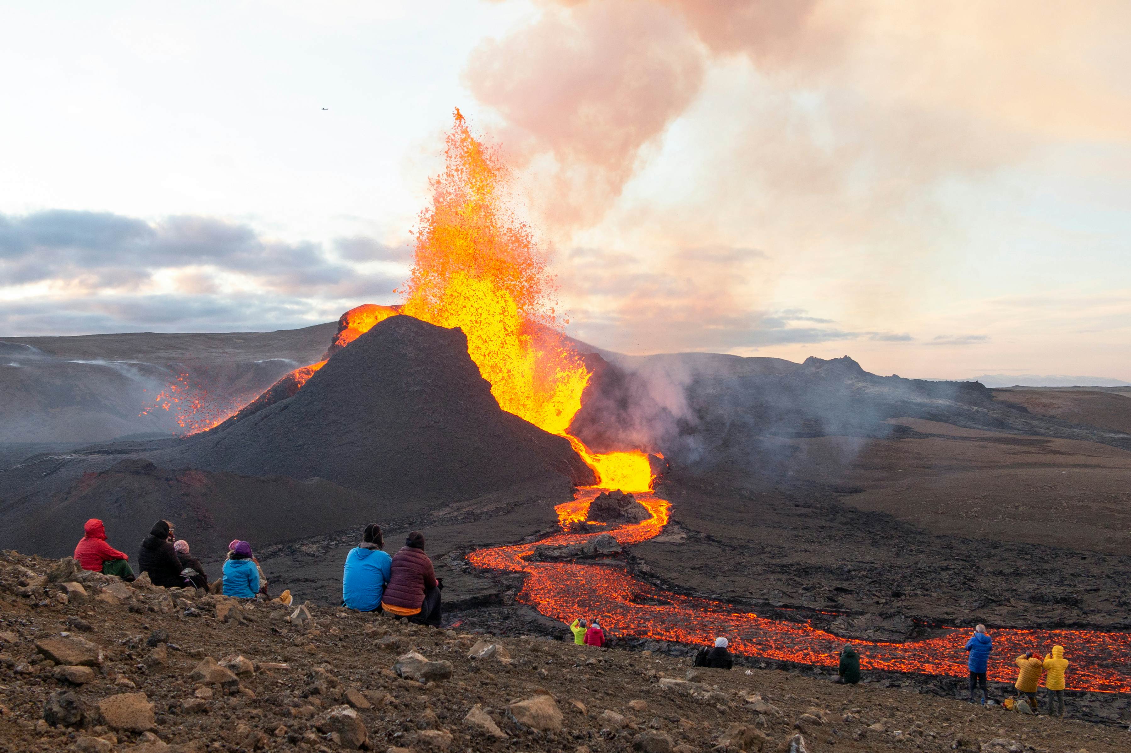 People sit on a rocky slope watching a volcano erupt from a safe distance. Lava explodes from the crater, and flows down in an orange ribbon.