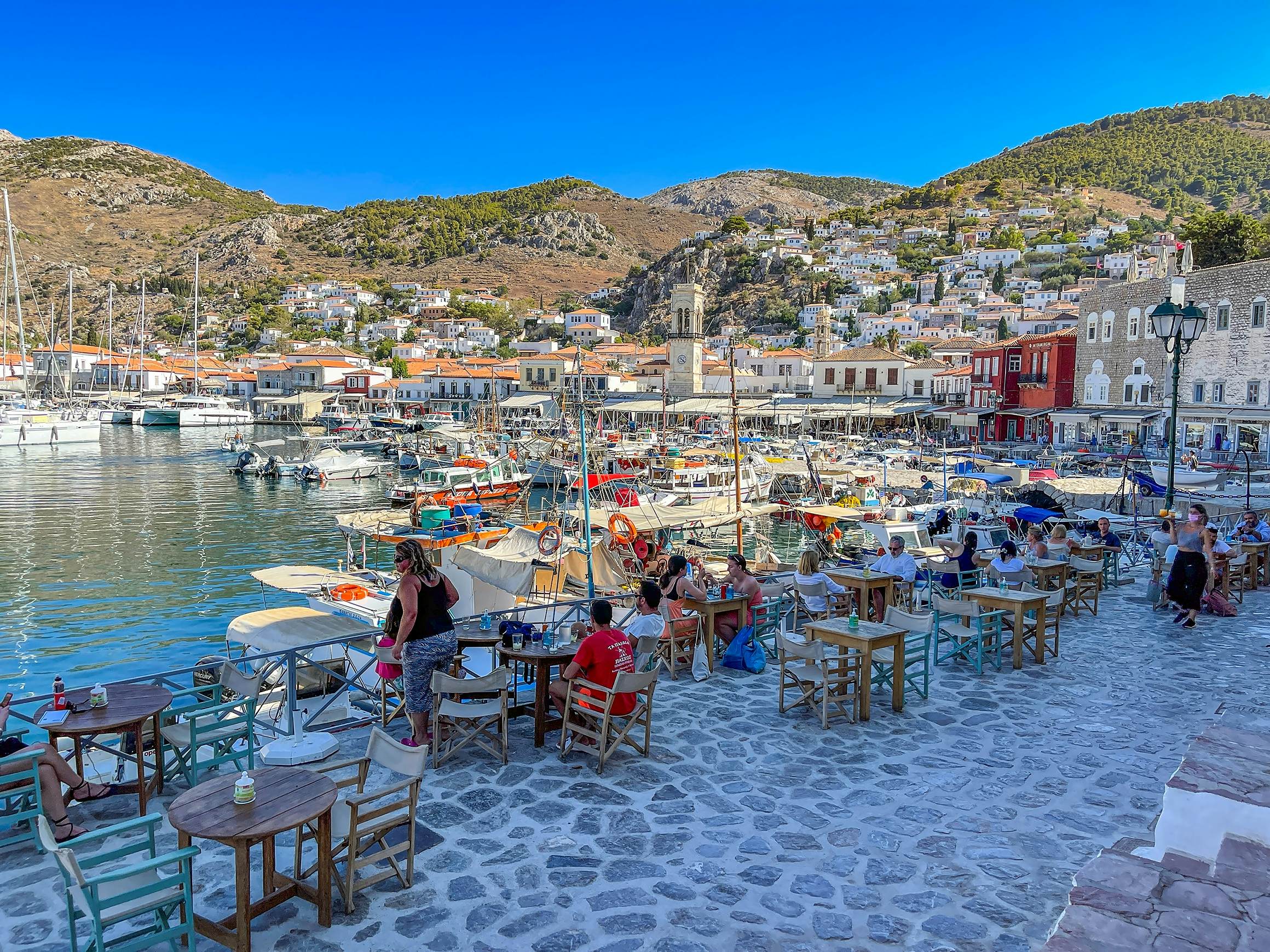 People sit at tables at an outdoor terrace overlooking the port of a village. Boats are moored in the water.