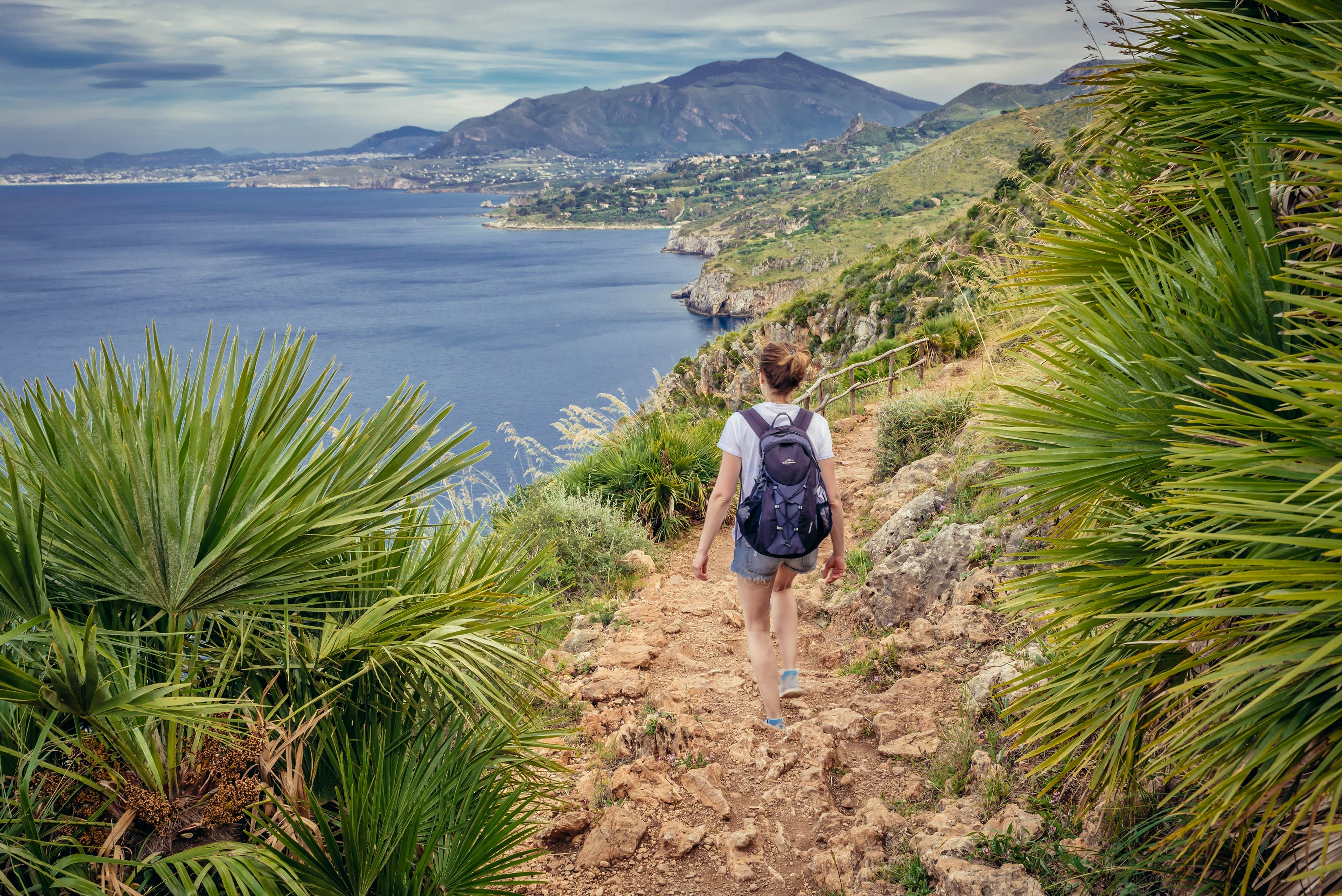 A woman is seen from behind walking along a dirt path on a hill by the seafront. Palm fronds and other vegetation frame the path.