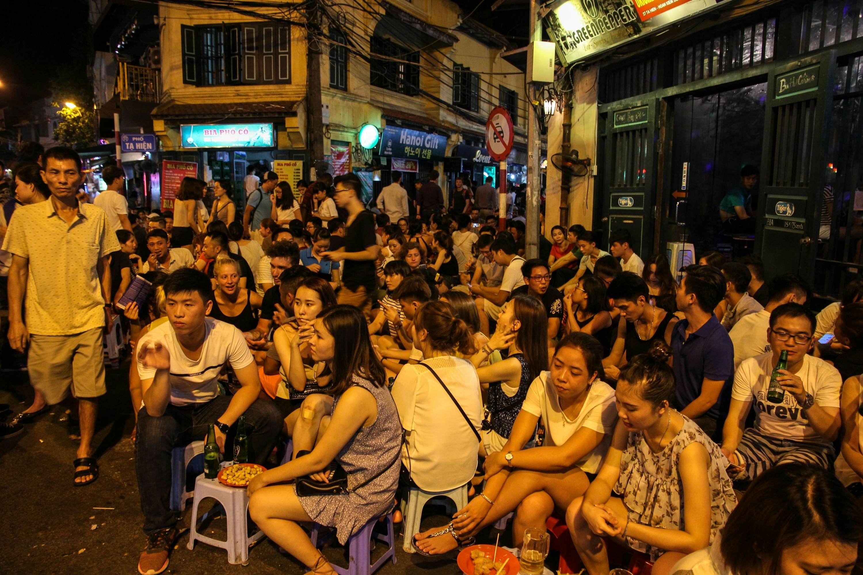 A crowded street corner in the old quarter of the city of Hanoi with young people enjoying bia hoi at small tables with plastic stools