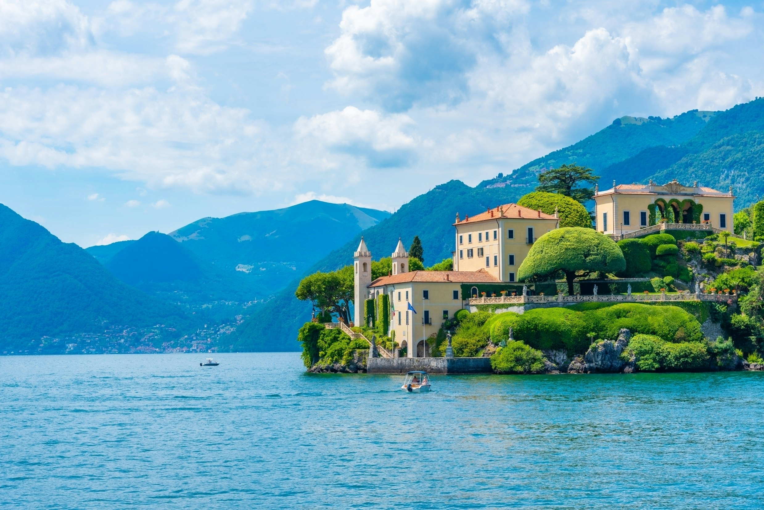 A lakeside villa with two pointed towers and large landscaped gardens, surrounded by hills.
